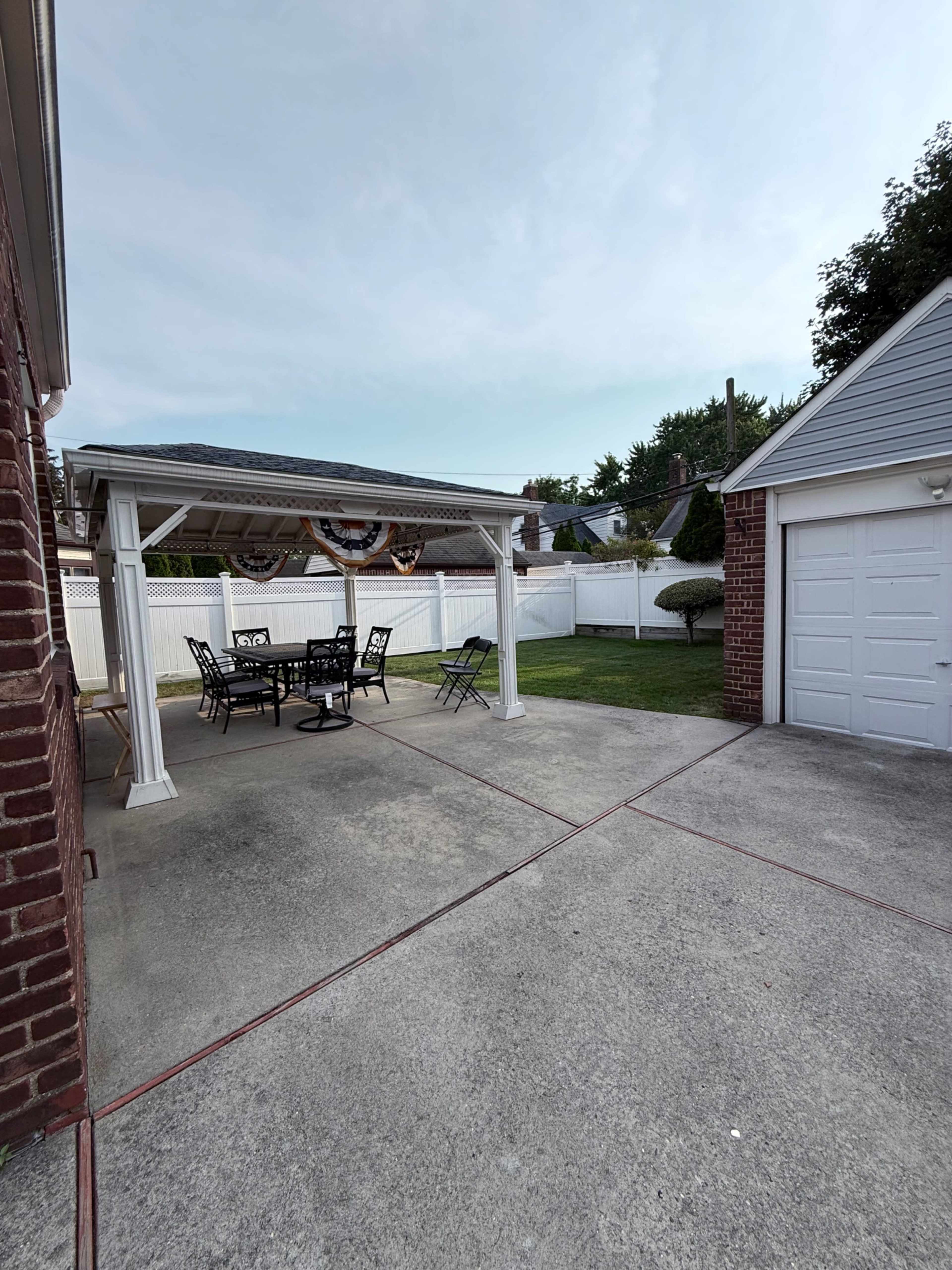 The image shows a covered patio with a black metal table and chairs, adjacent to a brick garage and enclosed by a white fence.