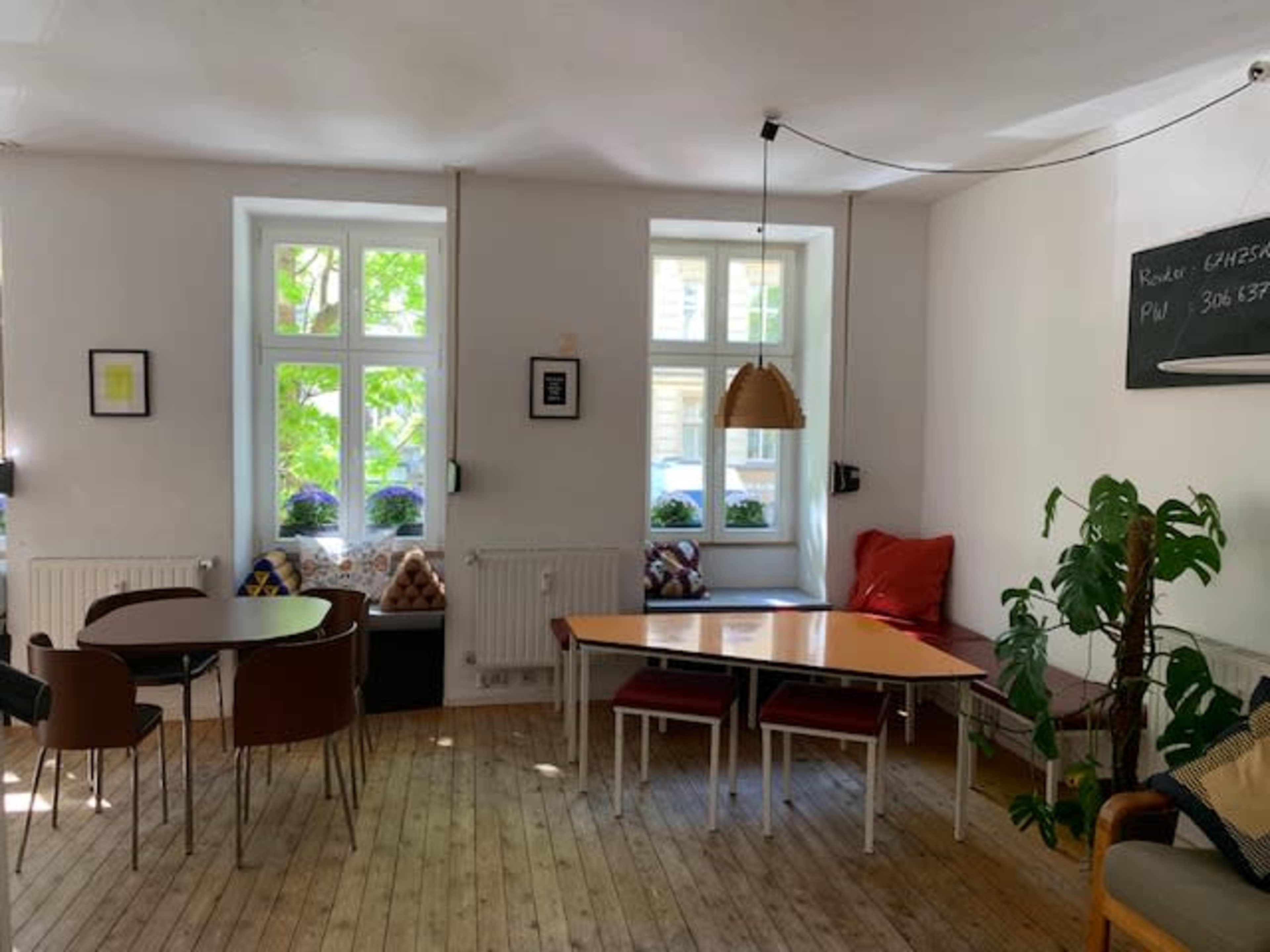 The image shows a bright, minimalist room with wooden floors, featuring a table and chairs, large windows, and a small seating area with cushions.