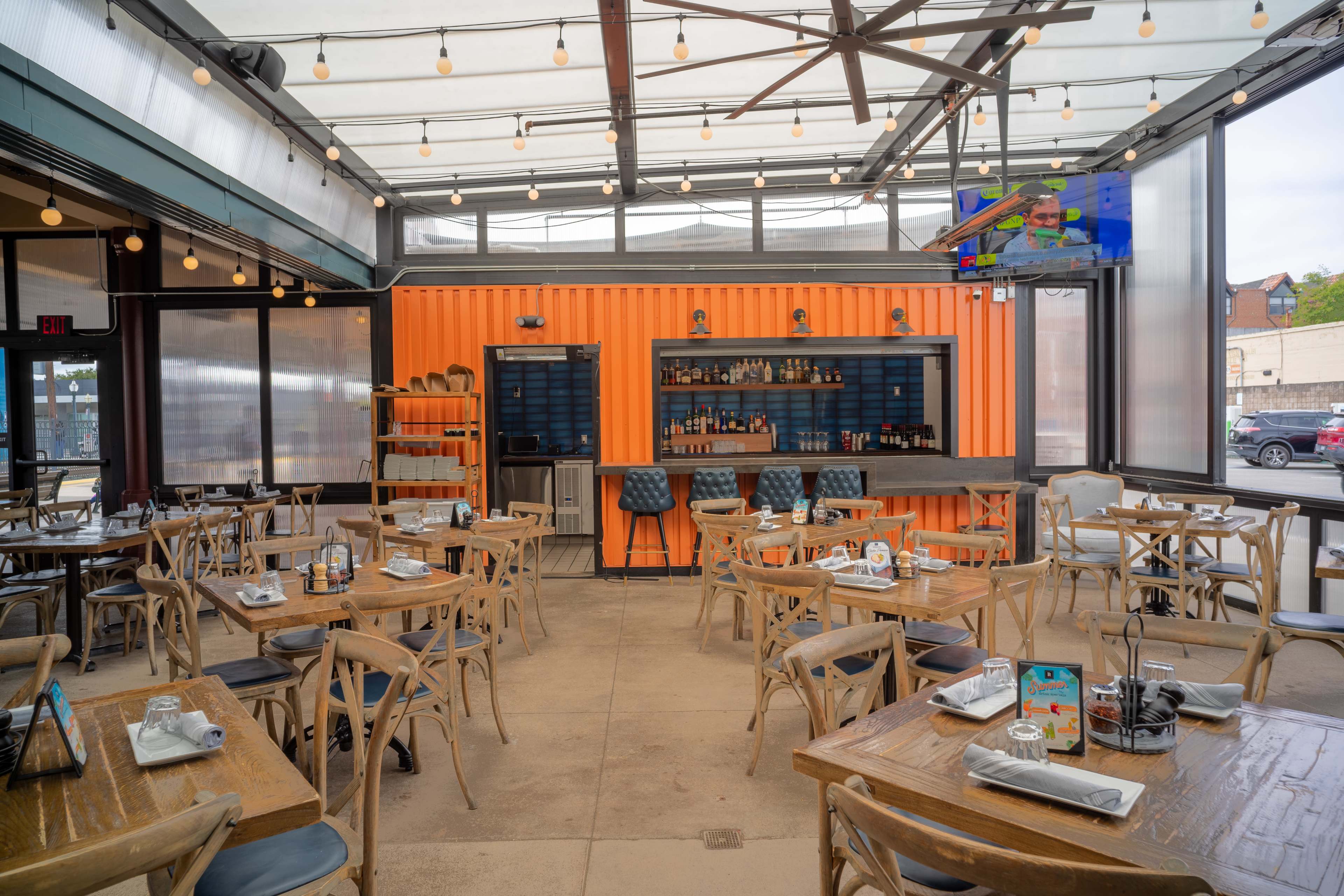 The image shows a spacious restaurant interior with wooden tables, chairs, and an orange shipping container wall serving as a backdrop for a bar area.