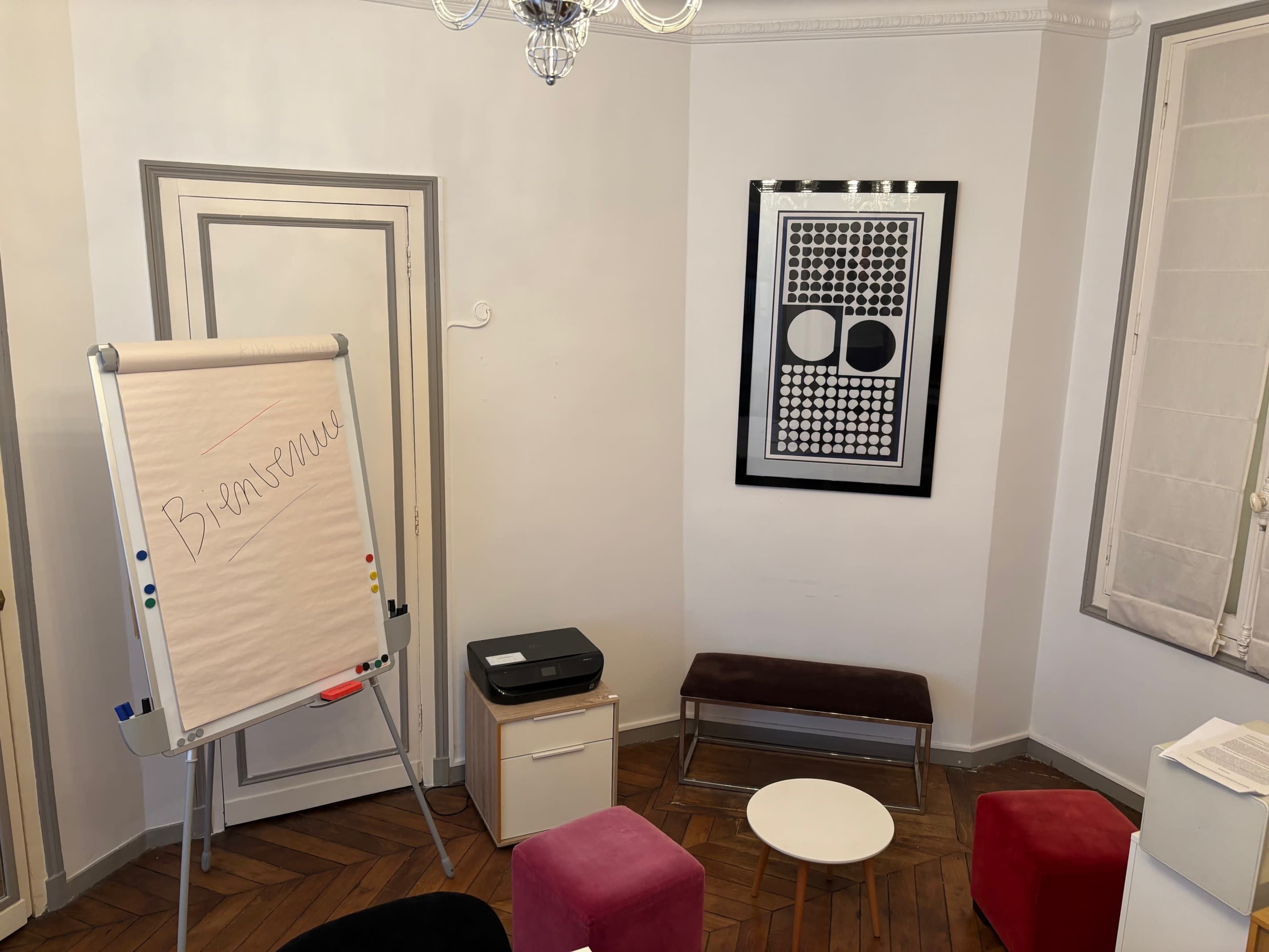 The image shows a small meeting room with a flip chart labeled "Bienvenue," a printer, a low table, and a decorative wall art piece.