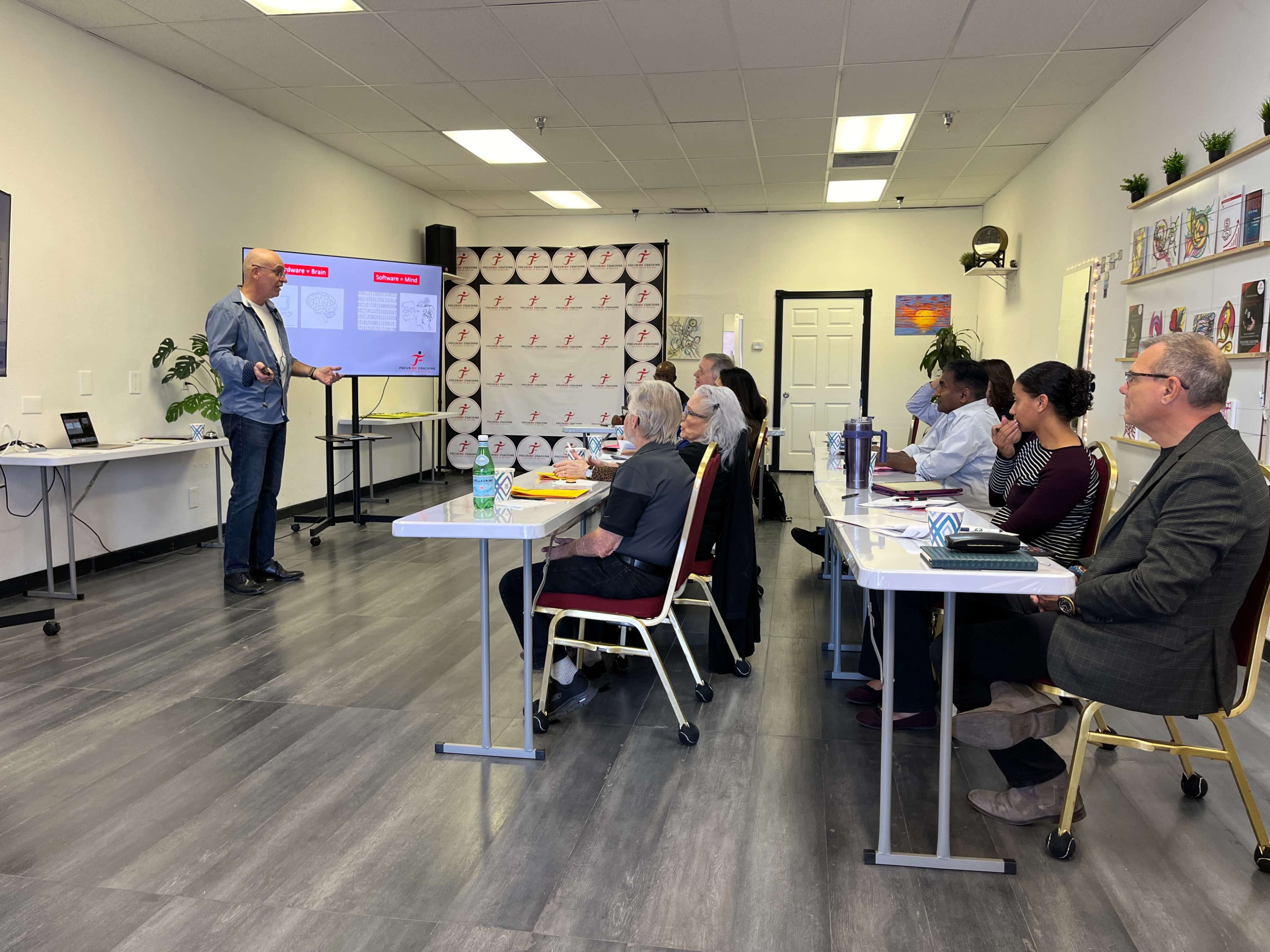 A speaker presents to a group of seated attendees in a classroom setting with a projector display on the wall.