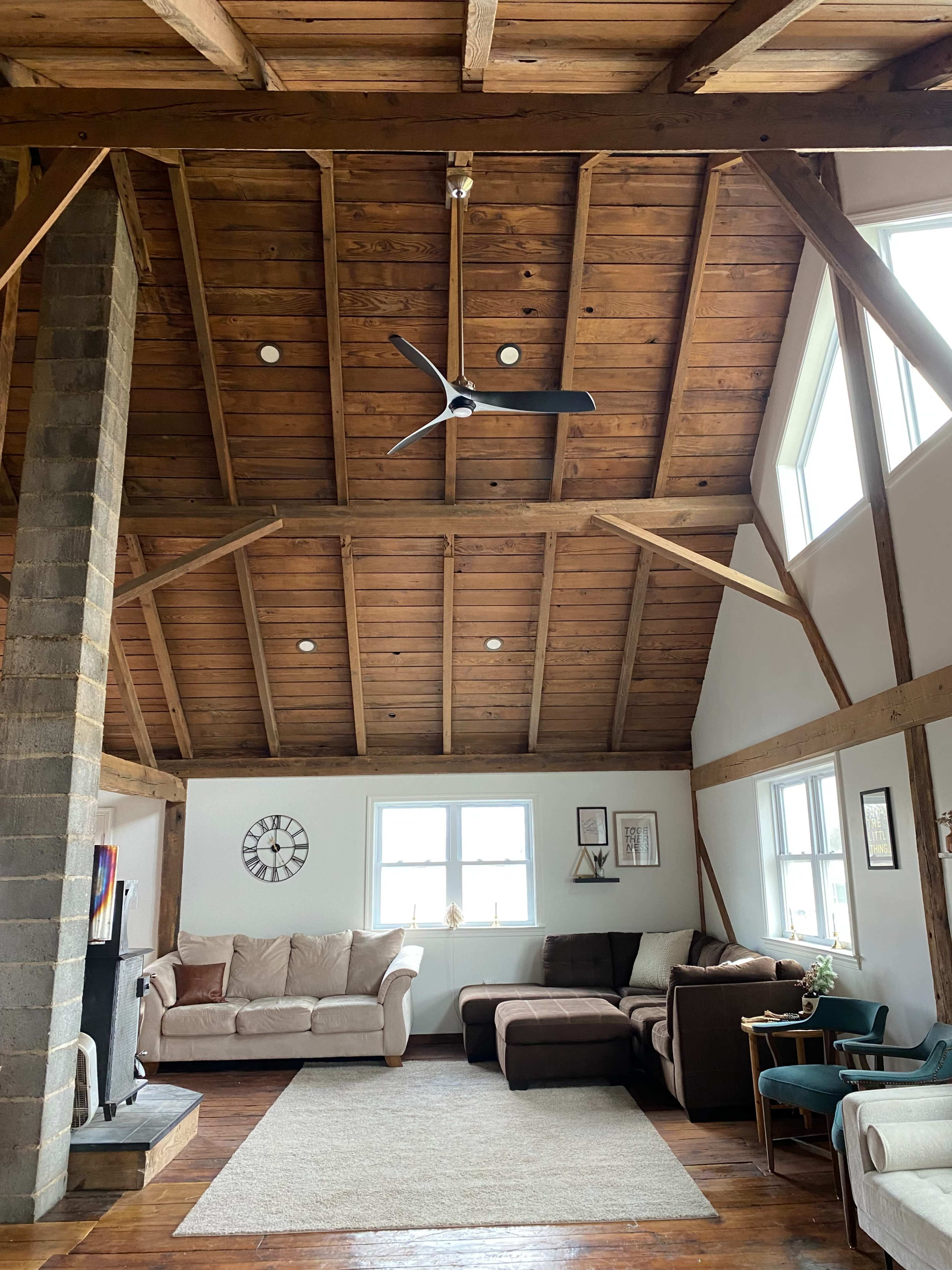 A spacious living room with a high, wooden ceiling, large windows, and a mix of beige and brown furniture arranged around a light-colored area rug.