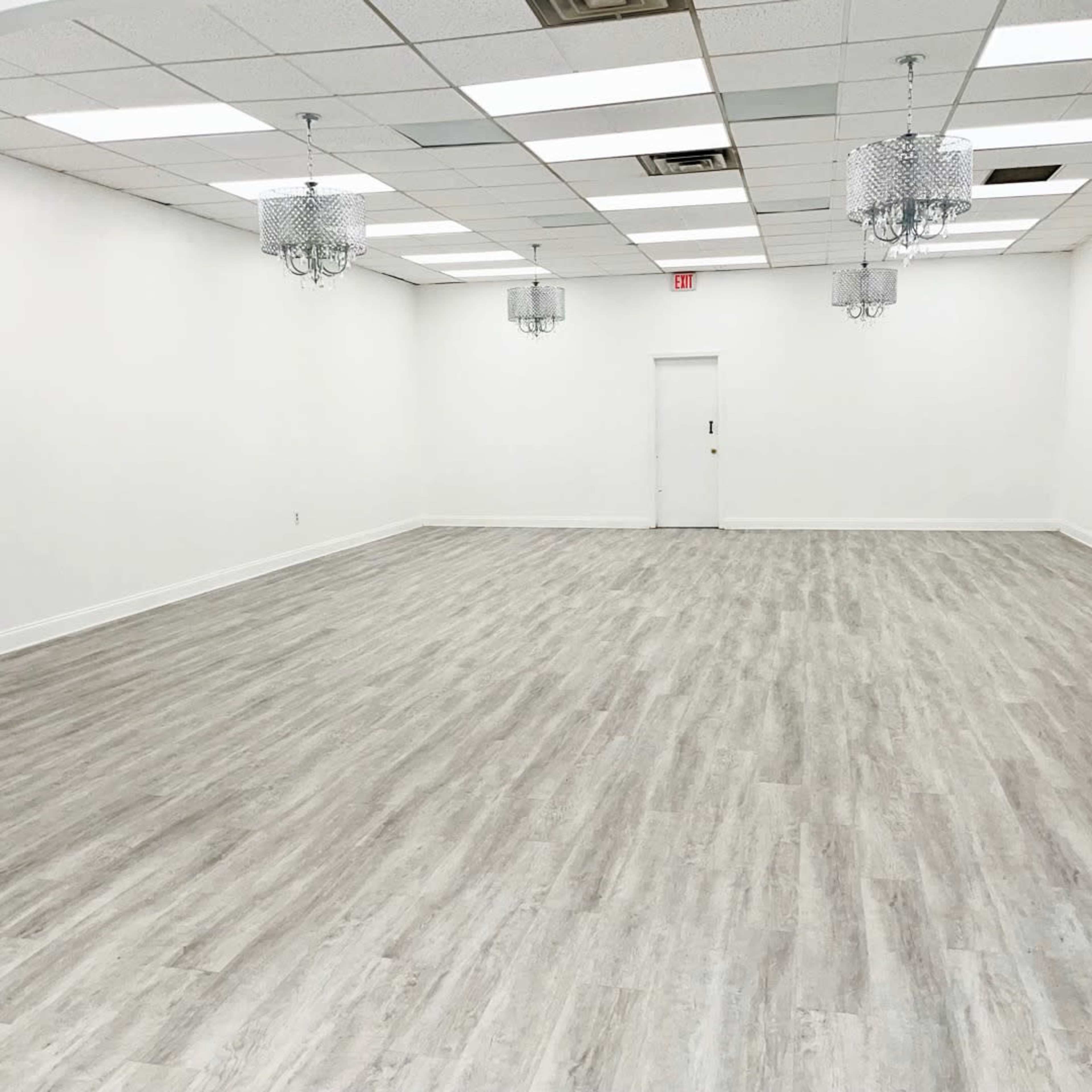 The image shows a spacious, empty room with light-colored wood flooring, white walls, and two decorative chandeliers hanging from the ceiling.