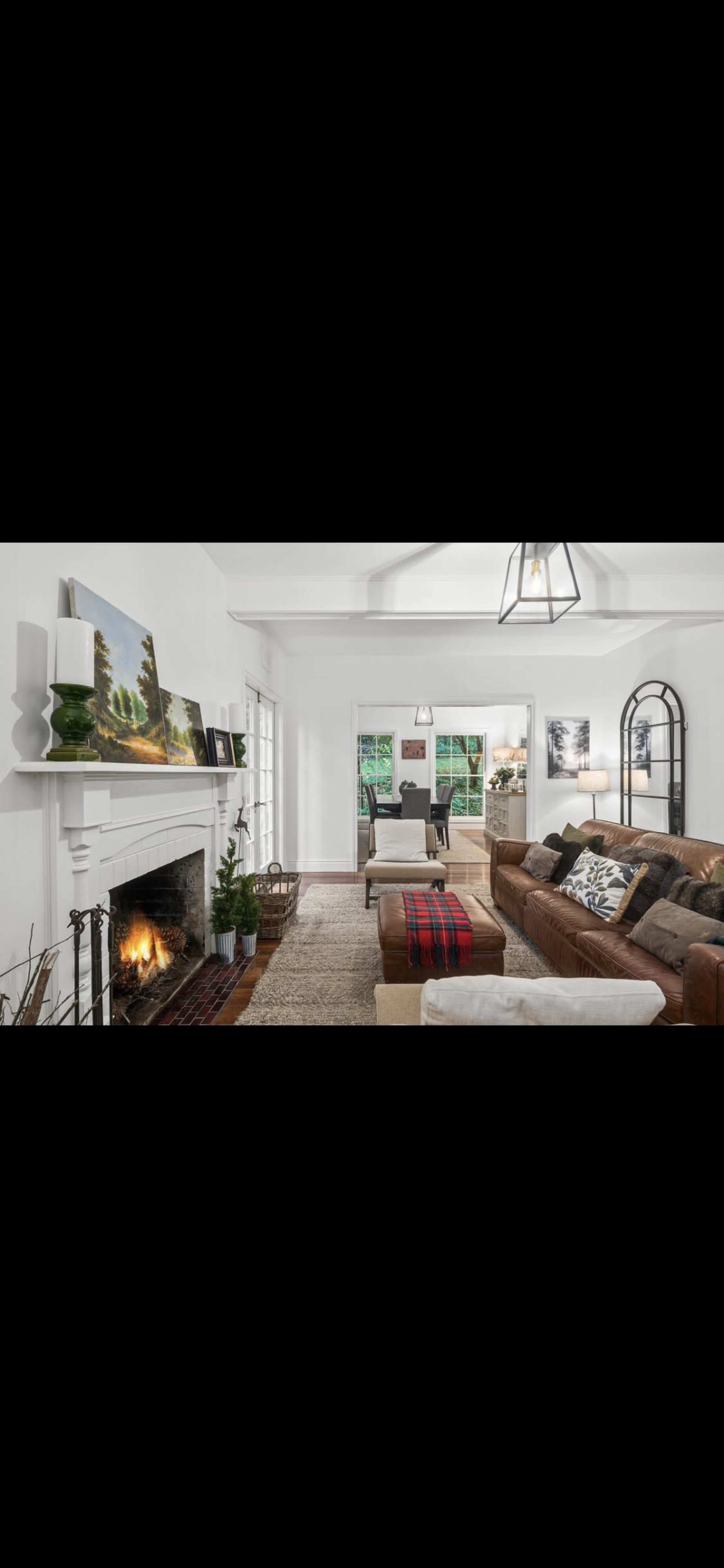 The living room features a brown leather sectional sofa, a coffee table with a red and black plaid throw, a fireplace with a white mantel, and large windows allowing natural light.