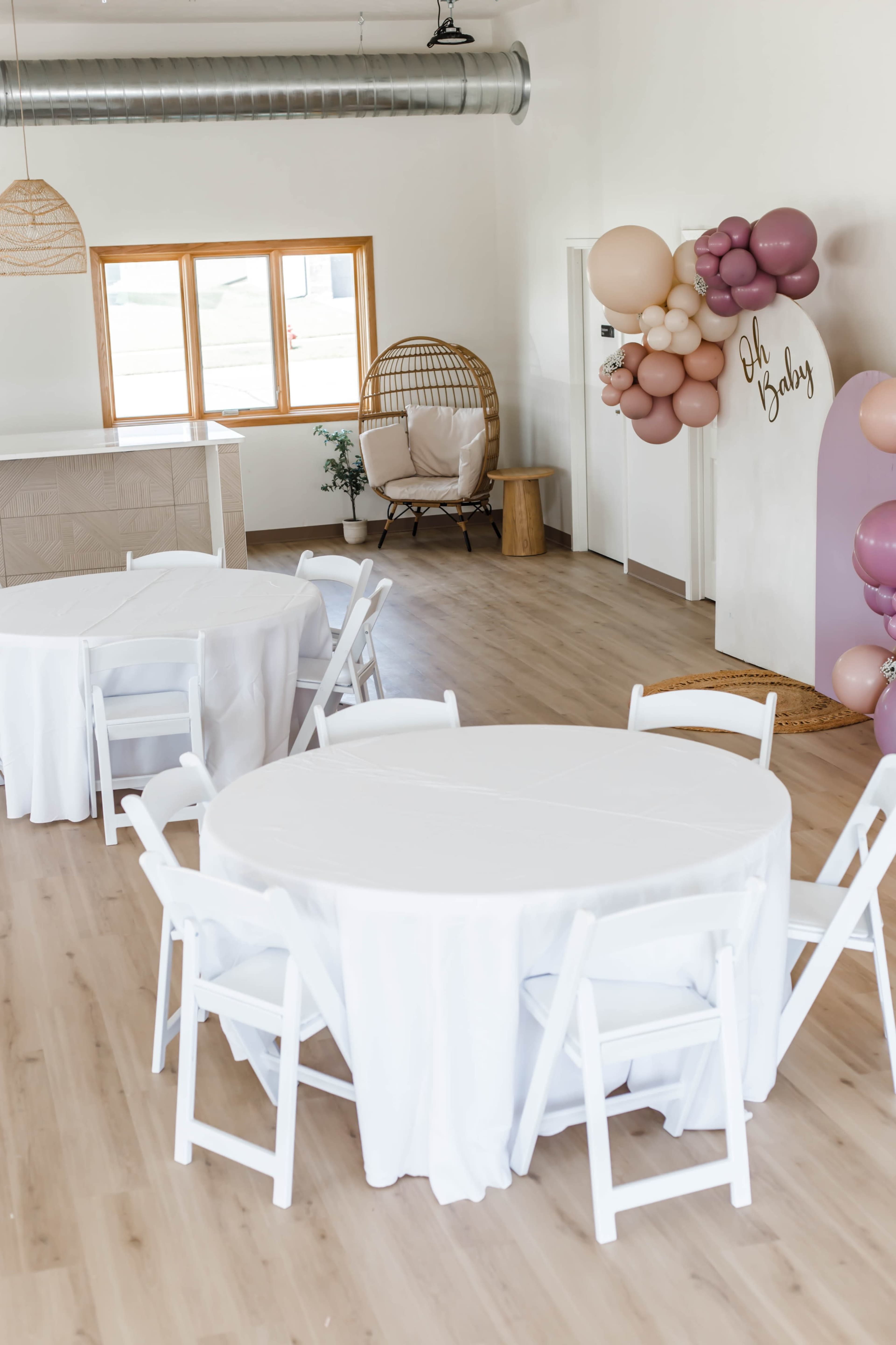 A modern event space with three round tables covered in white tablecloths, a decorative balloon arrangement, and a rattan chair in the corner.