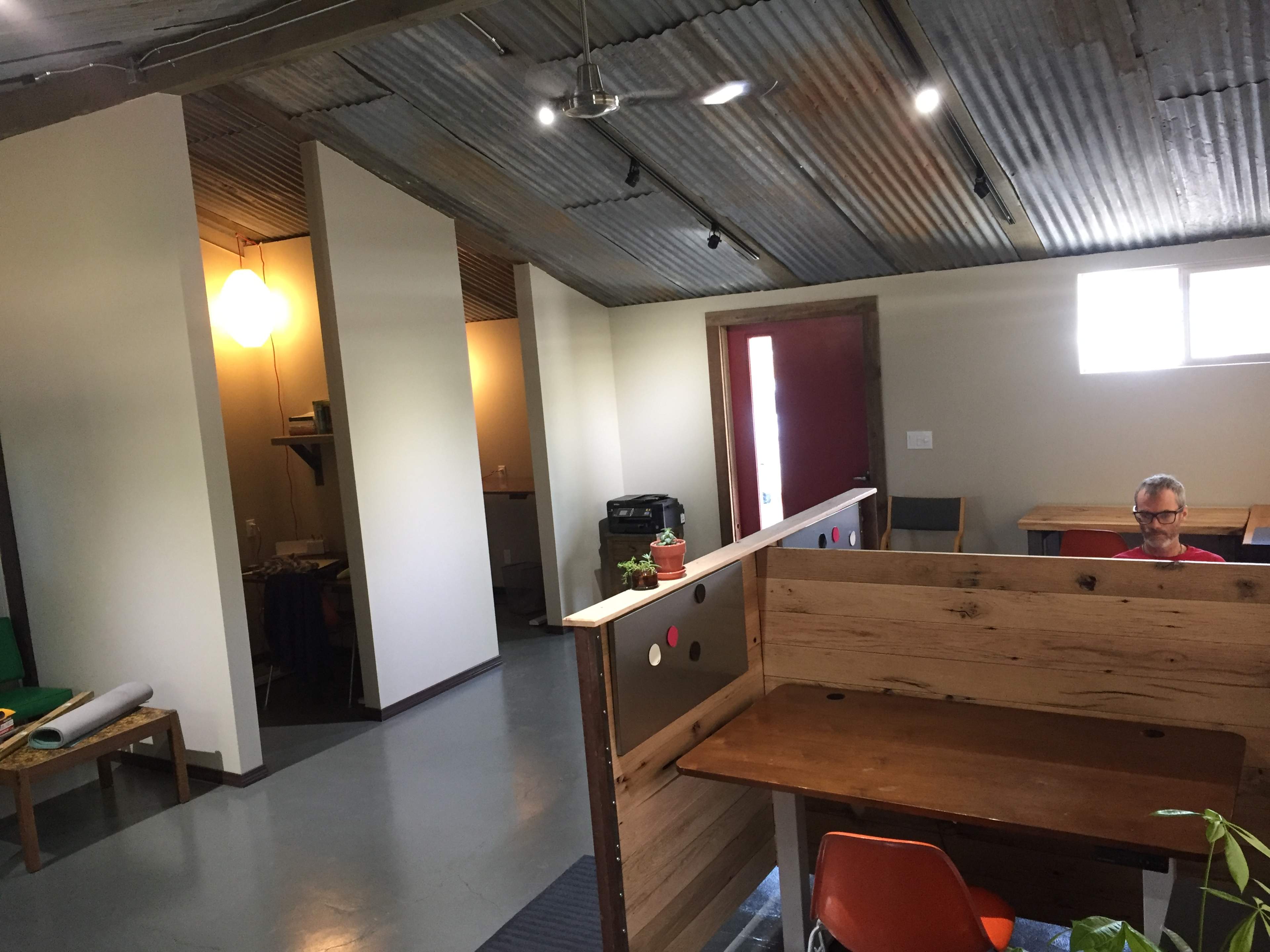 An open workspace features wooden desks, a small seating area, and a doorway leading to another room with a kitchenette.