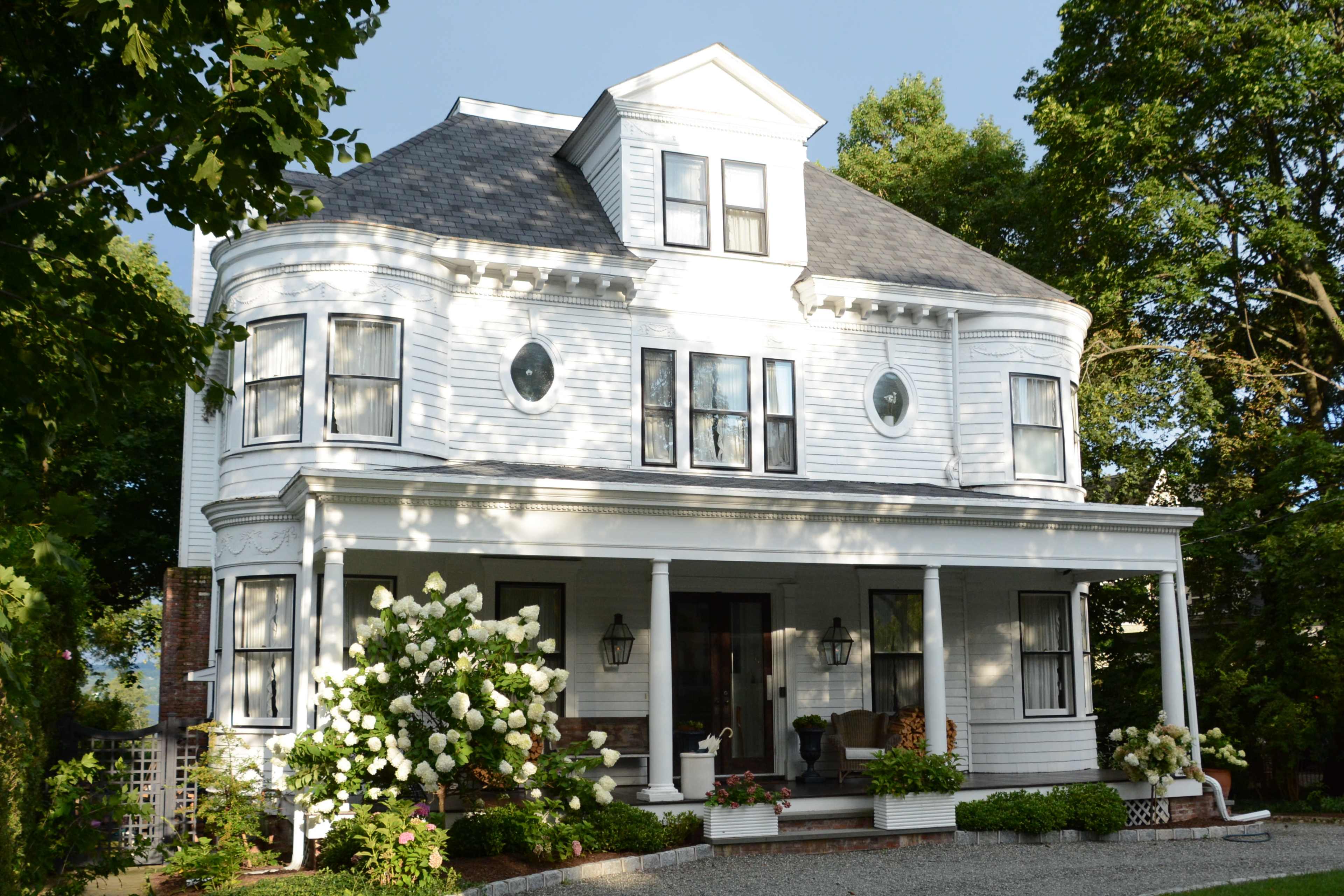 A two-story, white Victorian-style house with a rounded front and large windows, set in a landscaped yard with blooming white flowers.