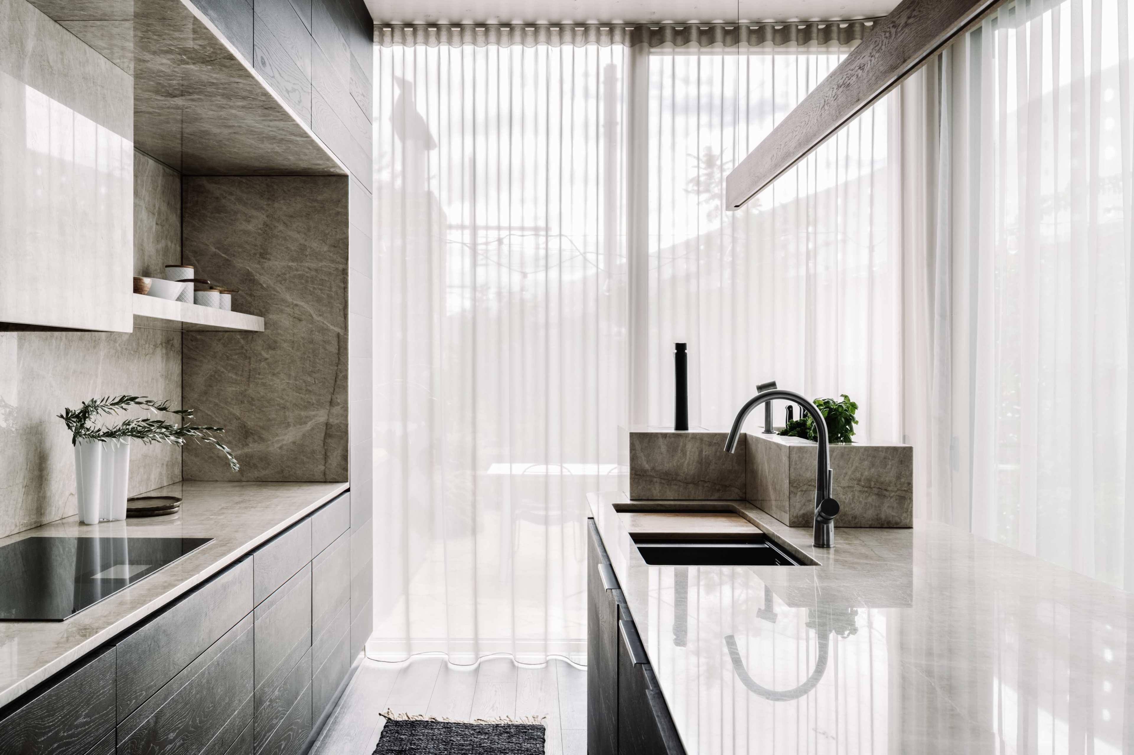 A modern kitchen features sleek dark cabinets, a stone countertop, and large windows with sheer curtains allowing natural light to fill the space.