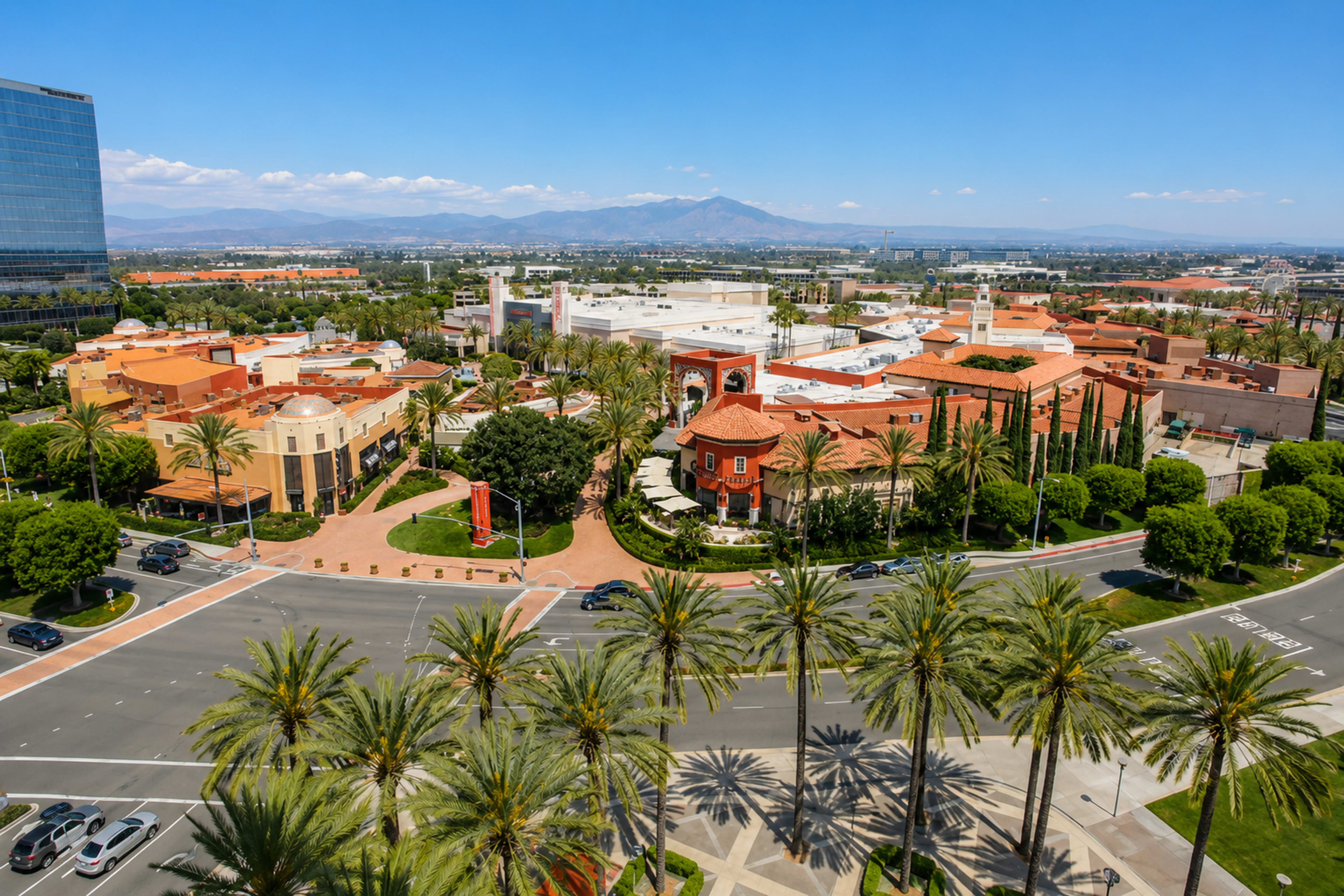 The image shows a sprawling urban area with palm trees lining the streets, featuring a mix of buildings and green spaces, set against a backdrop of mountains.