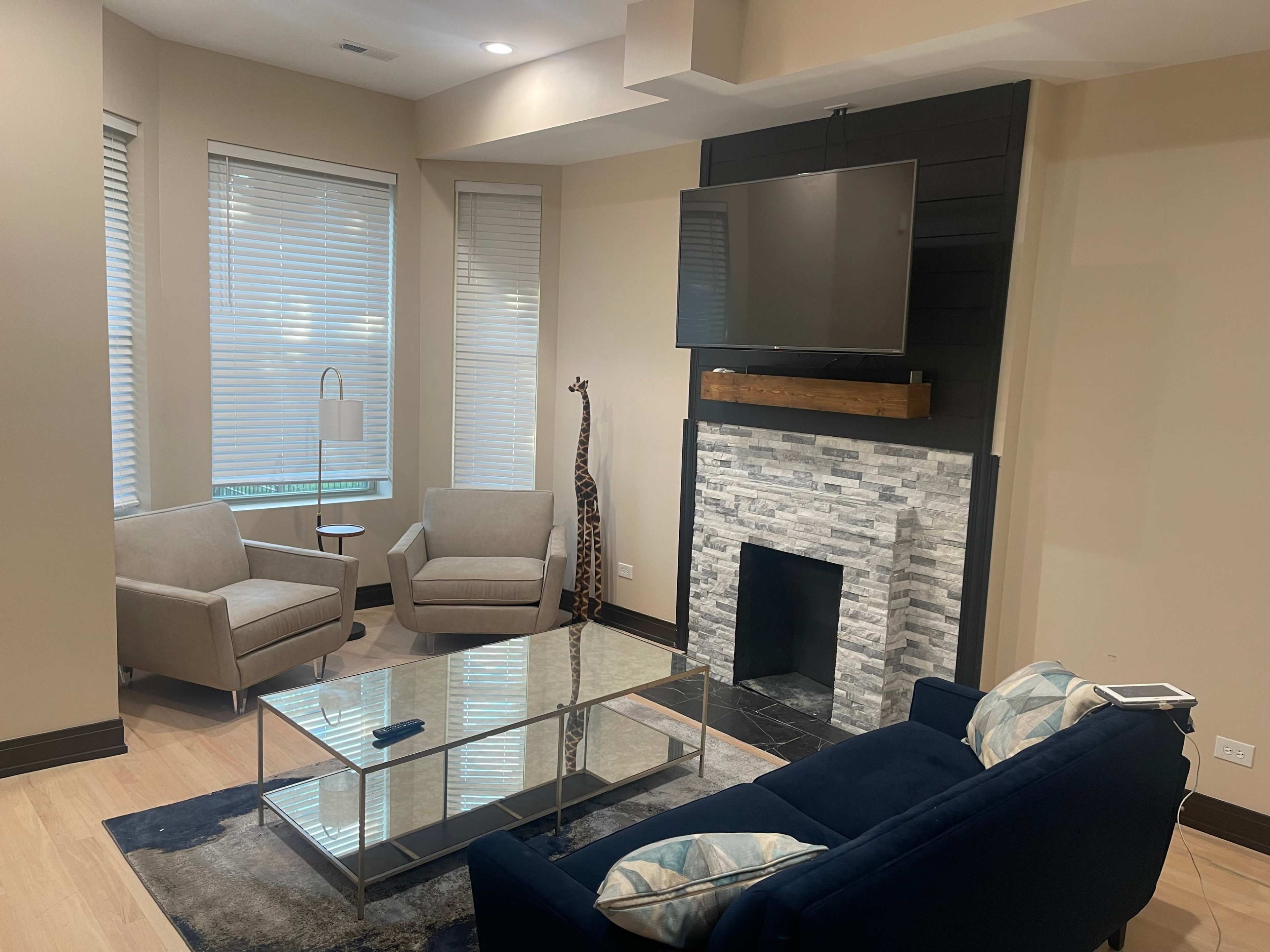 A modern living room with two upholstered chairs, a blue sofa, a glass coffee table, a stone fireplace, and a mounted television.