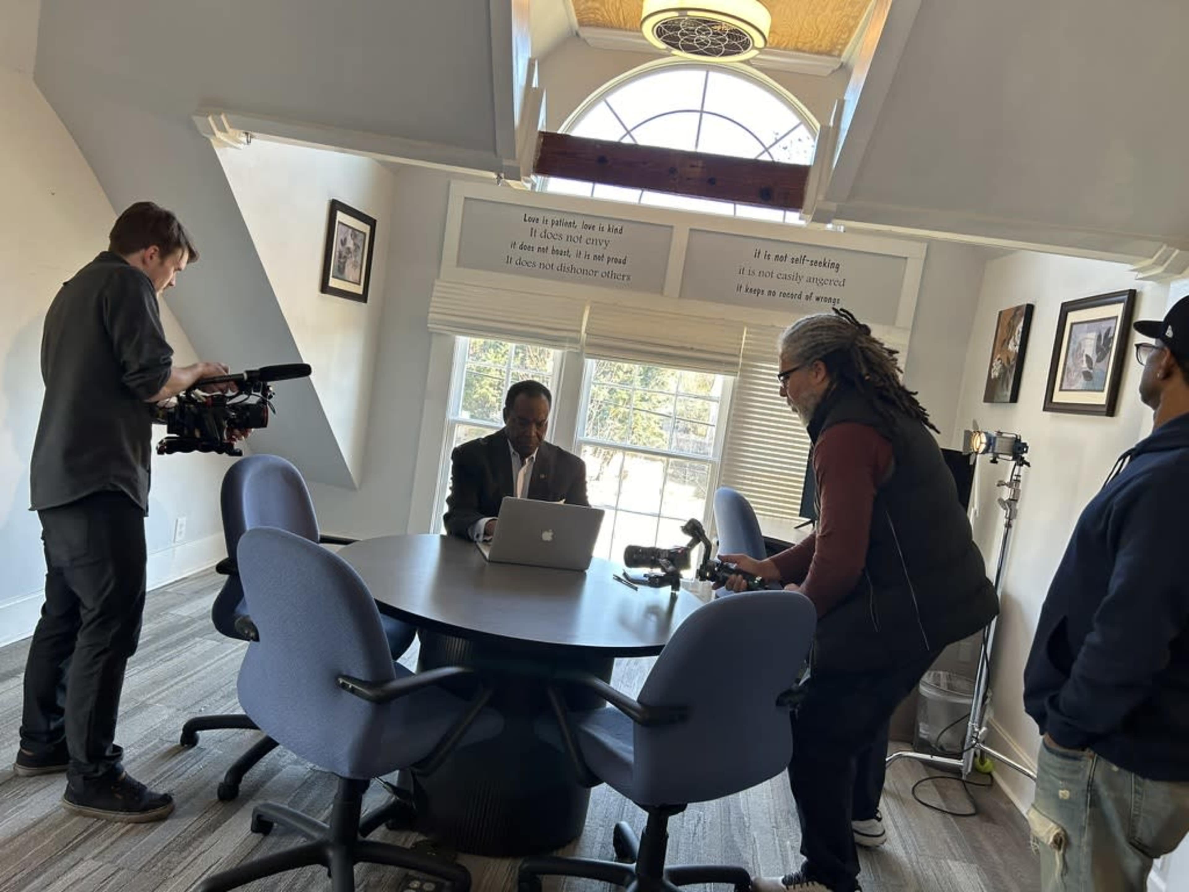 A group of three people is filming a man sitting at a round table with a laptop in a well-lit office space.