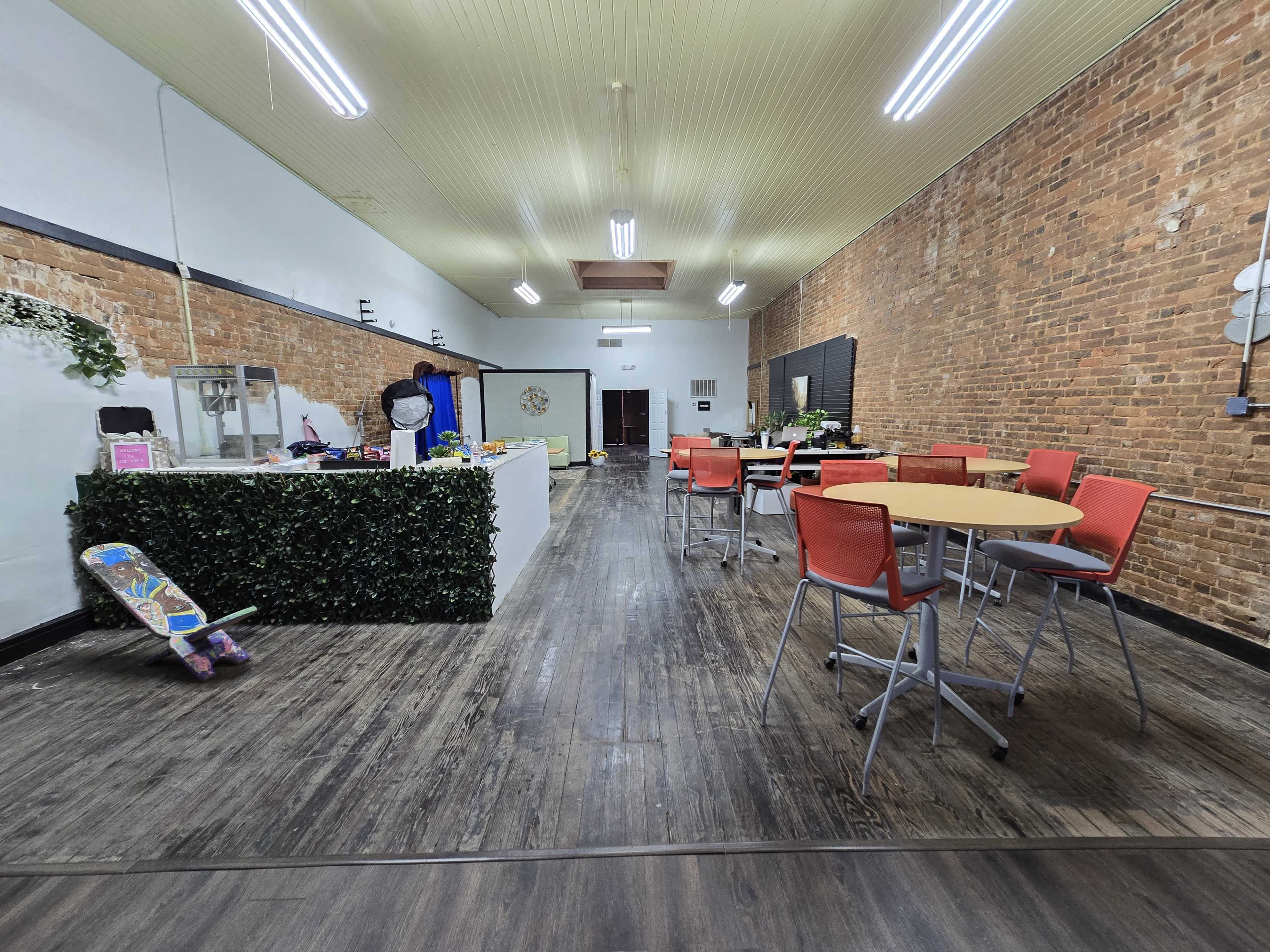 The image shows a spacious indoor area with exposed brick walls, featuring tables and chairs arranged for seating, along with a service counter at one end.