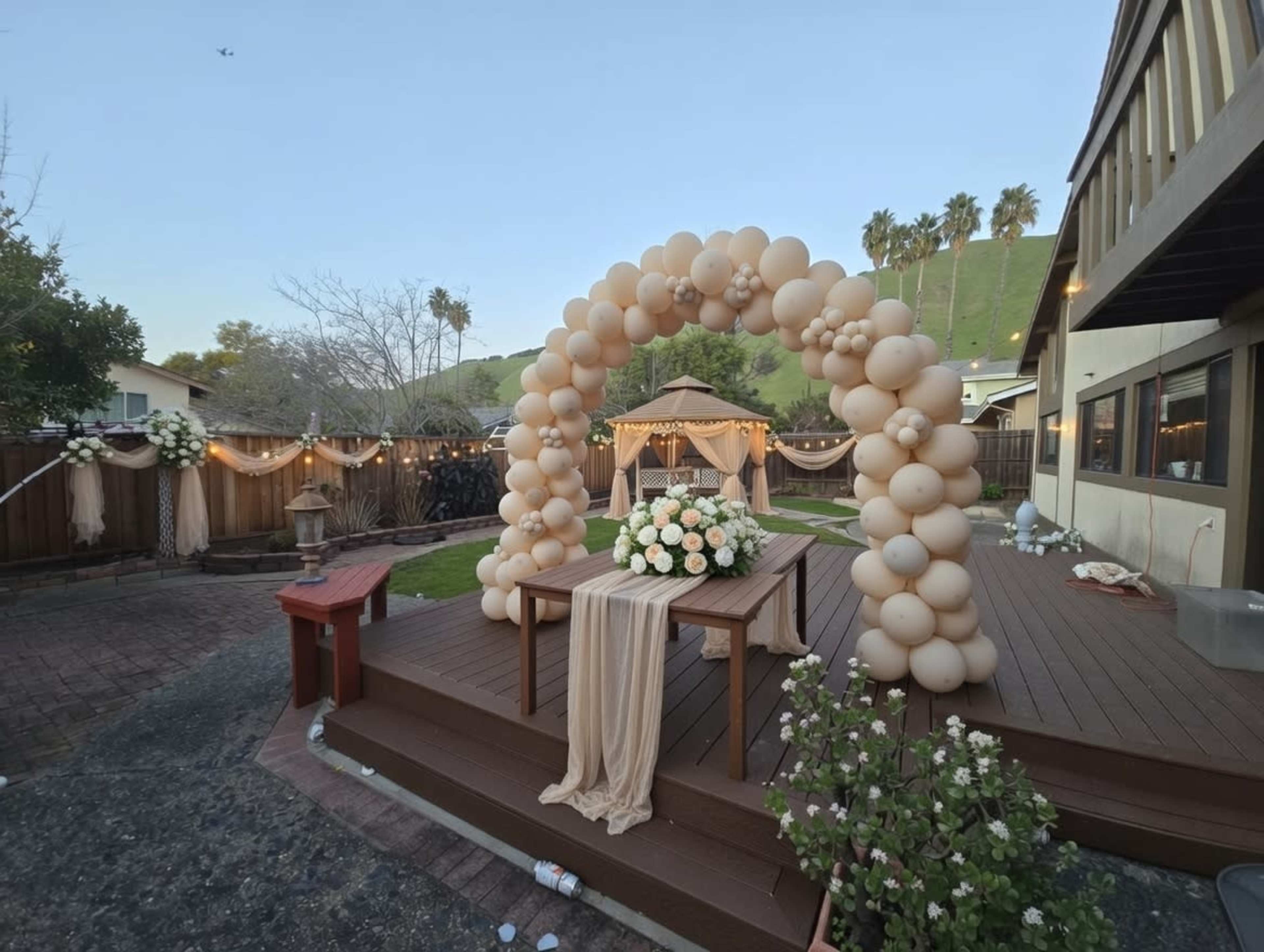 The image shows a decorated outdoor space for an event, featuring a balloon arch, a table with flowers, and a gazebo amidst a backdrop of greenery.