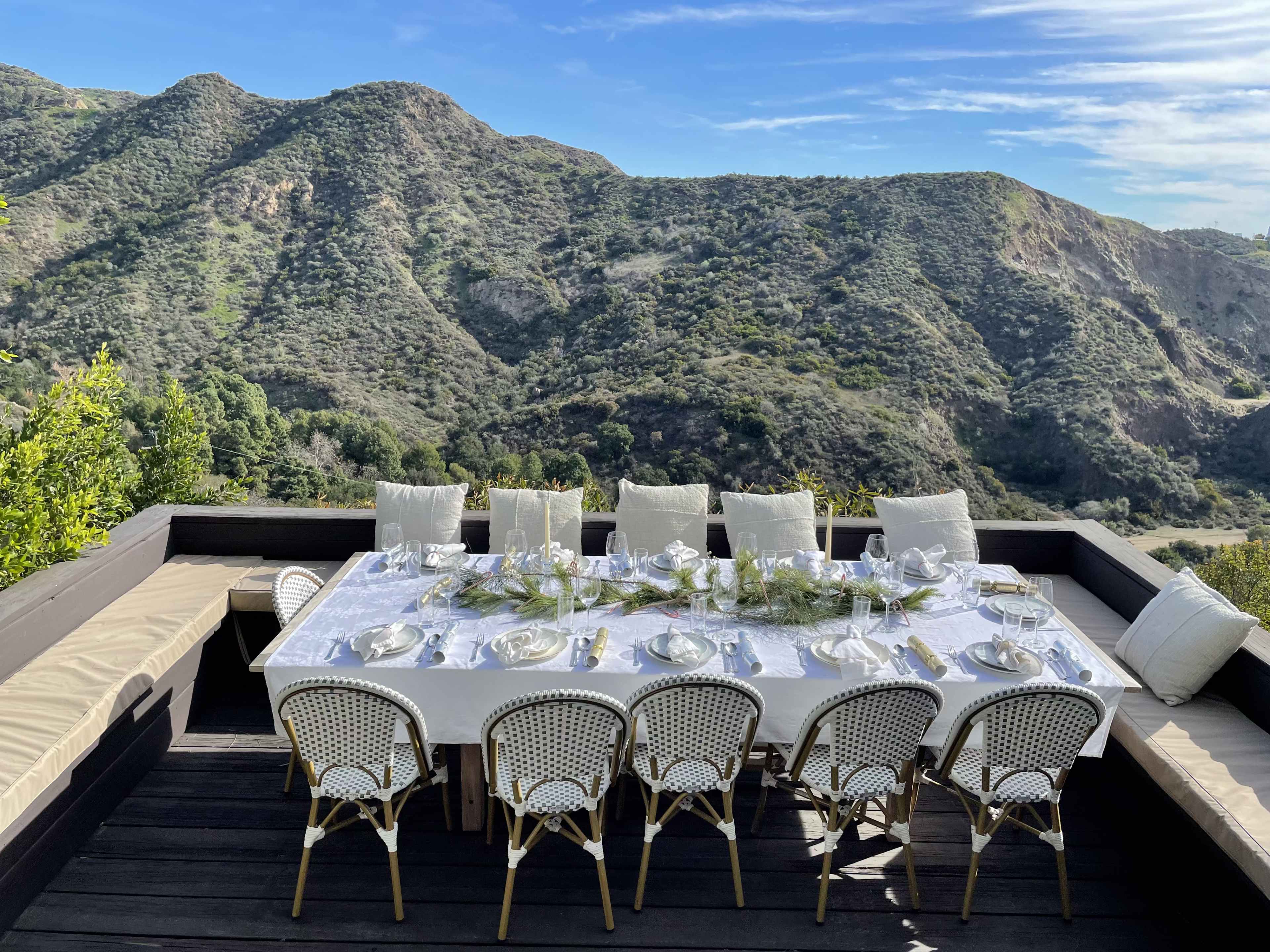 A large dining table set for a meal is positioned on a deck overlooking lush green hills.