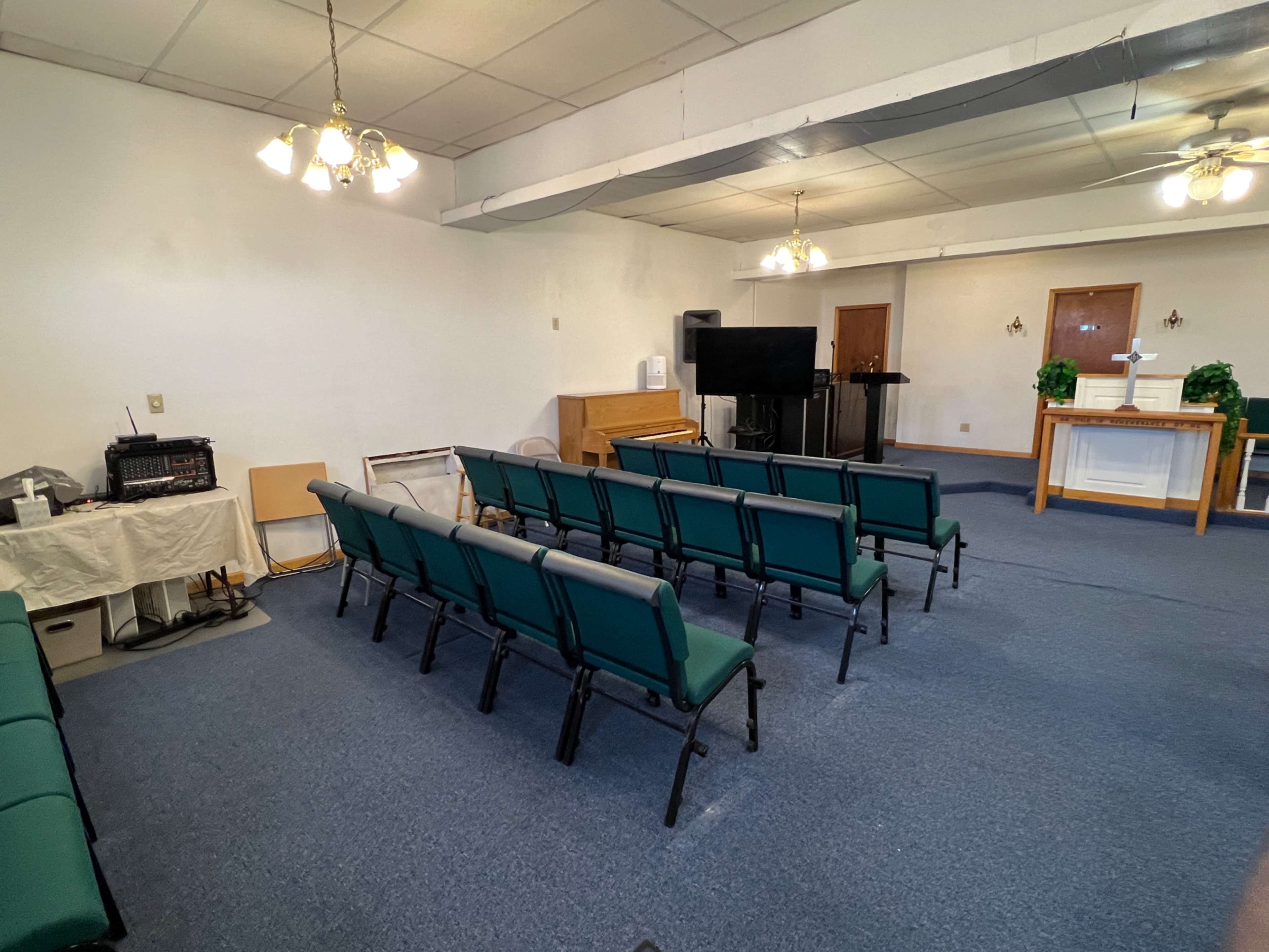 The image shows a sparsely furnished room with several rows of green chairs facing a low podium and a television screen.