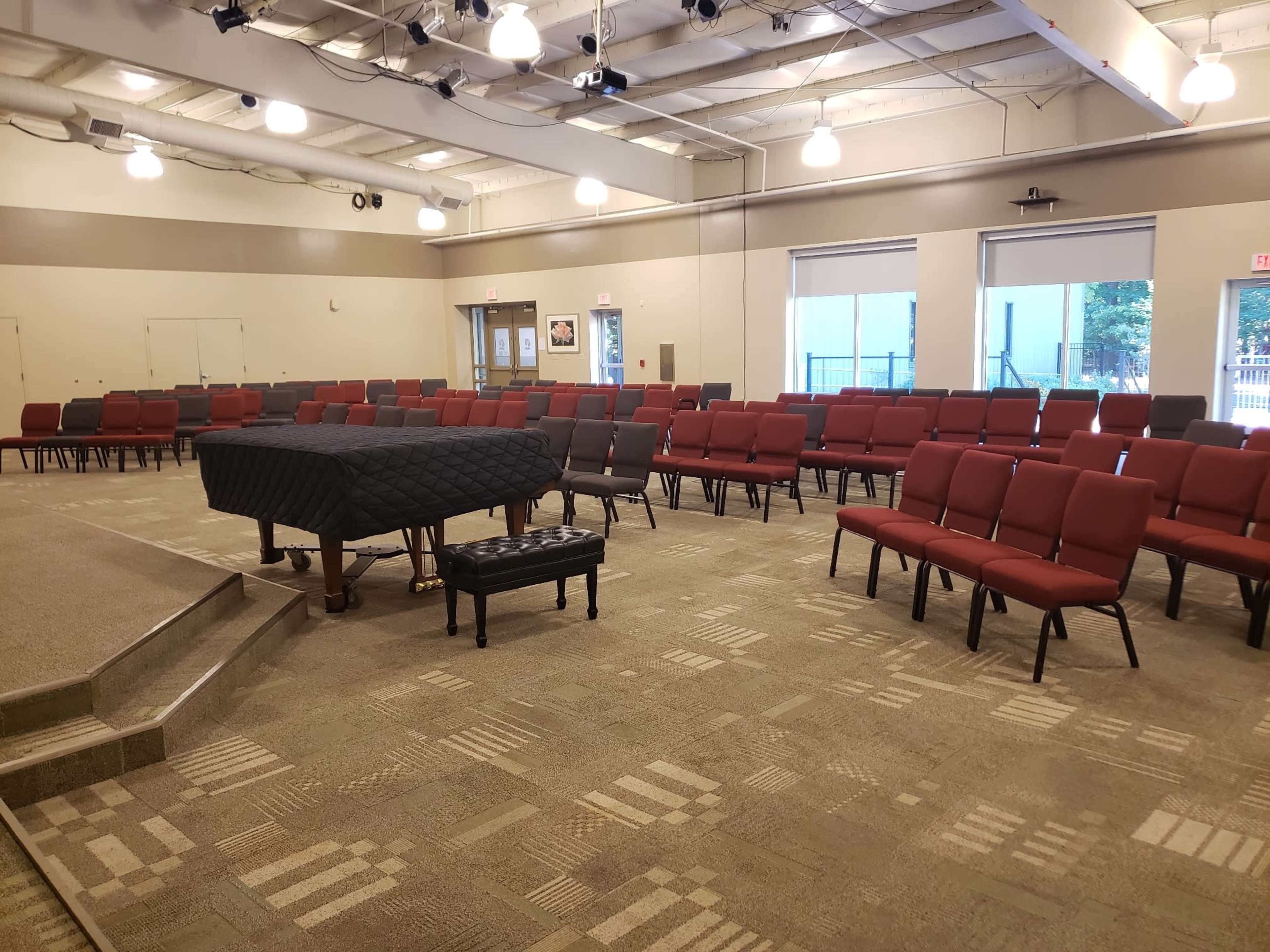 A room with red chairs arranged in rows facing a black grand piano on a stage, with natural light illuminating the space from large windows.