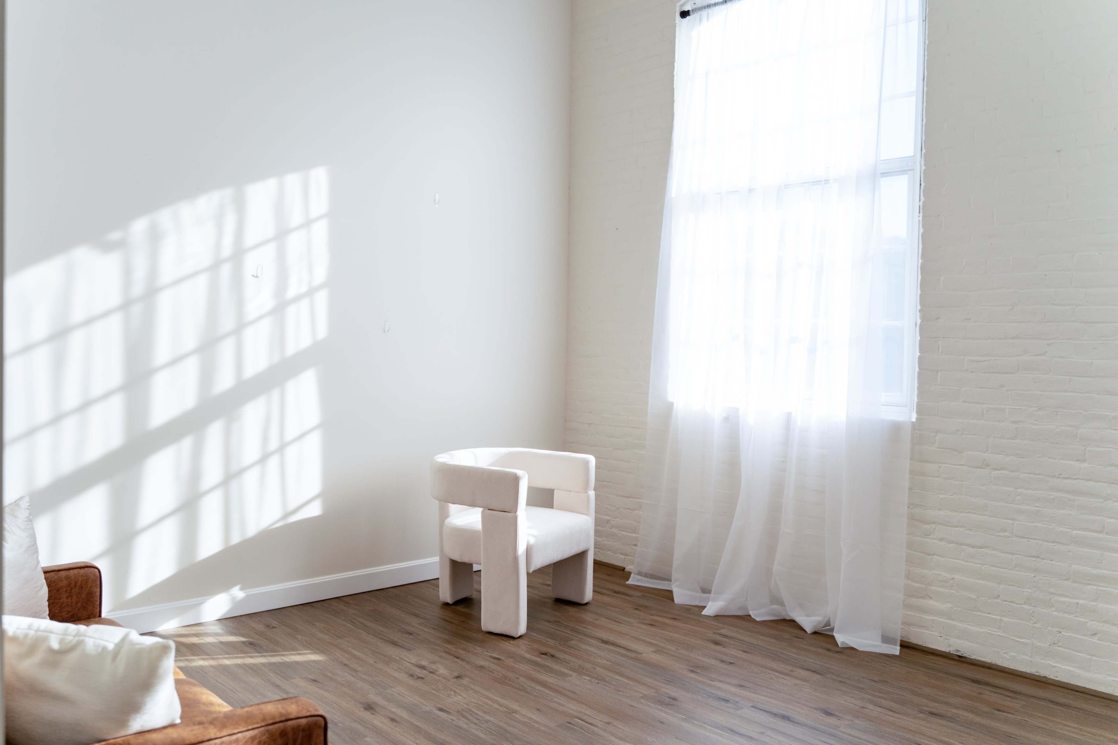 A minimalistic room features a white chair beside a window with sheer curtains allowing light to cast shadows on the floor.