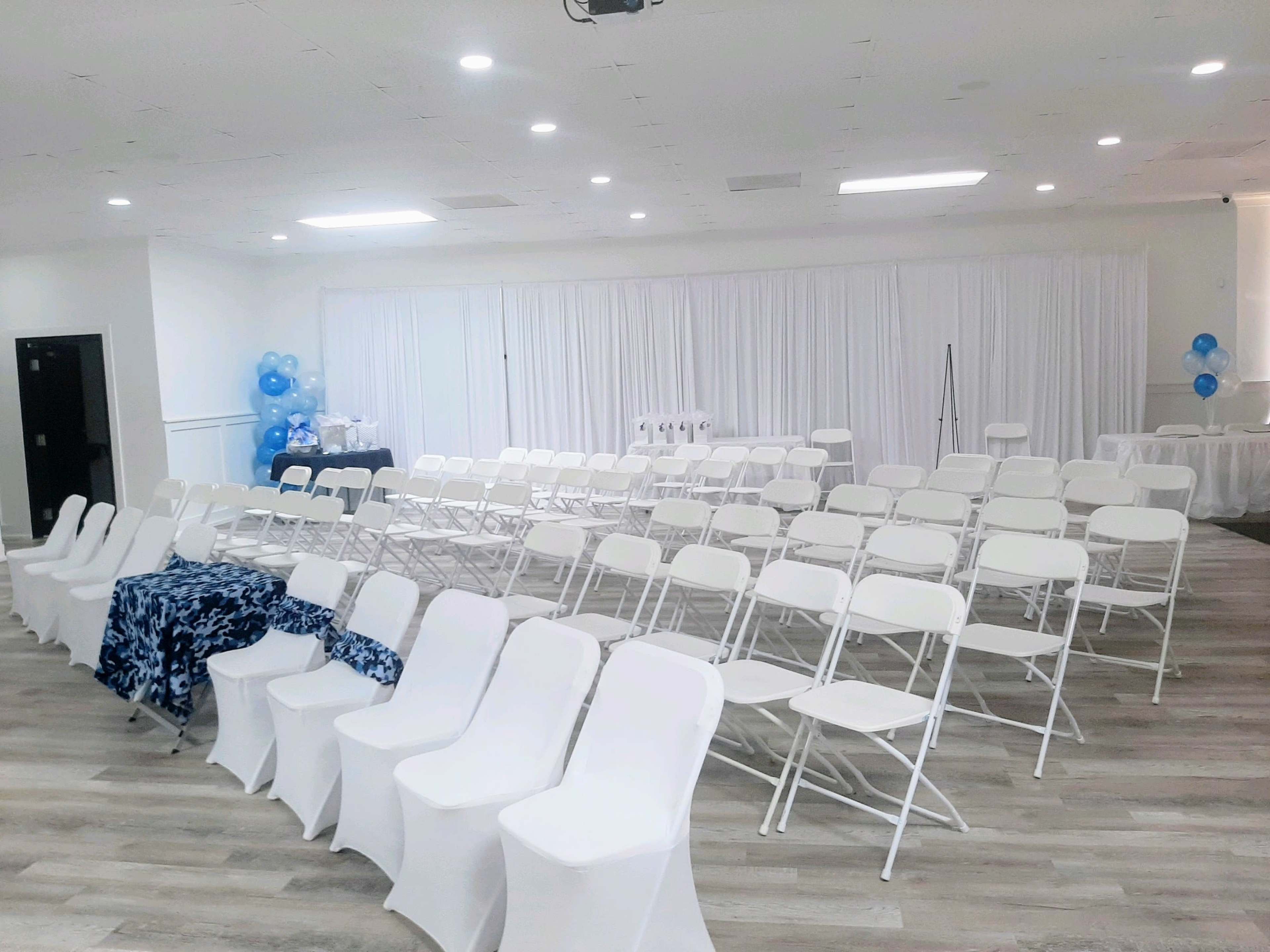A spacious event venue set up with rows of white folding chairs facing a white curtain backdrop, along with blue decorations and tables.