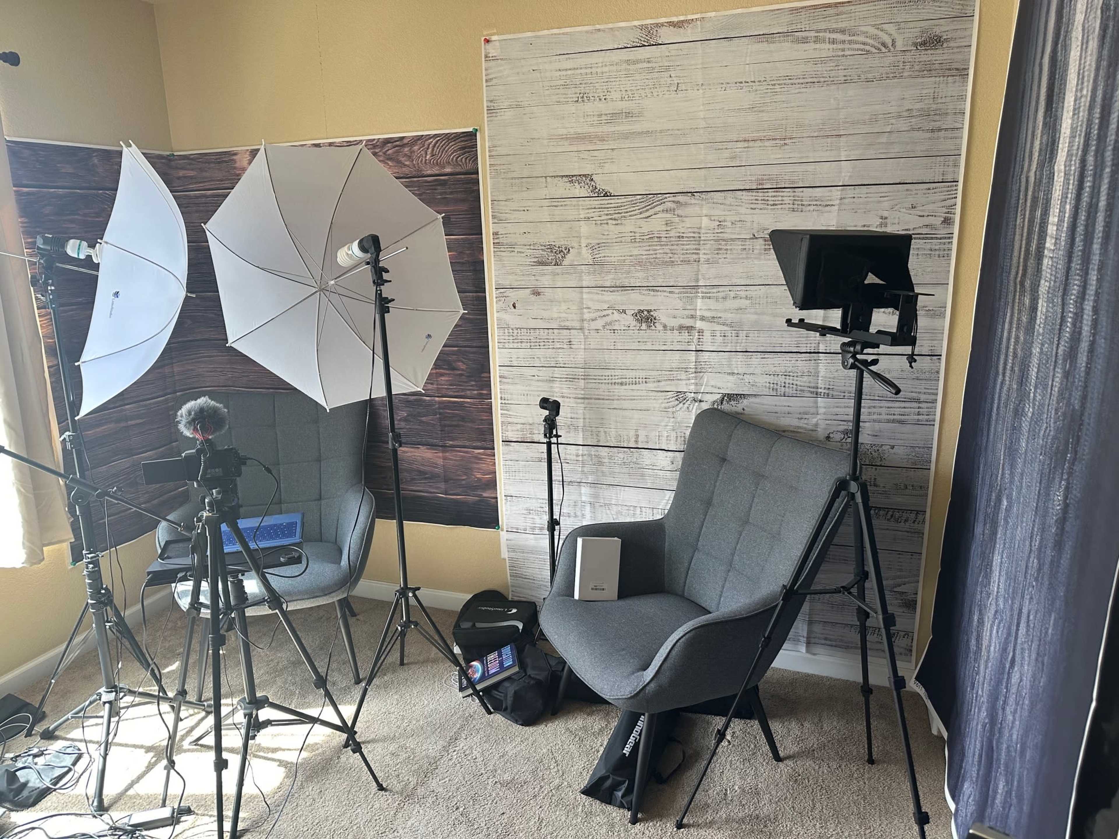 A studio setup featuring softbox lights, a camera on a tripod, a gray chair, and a backdrop resembling wooden planks.