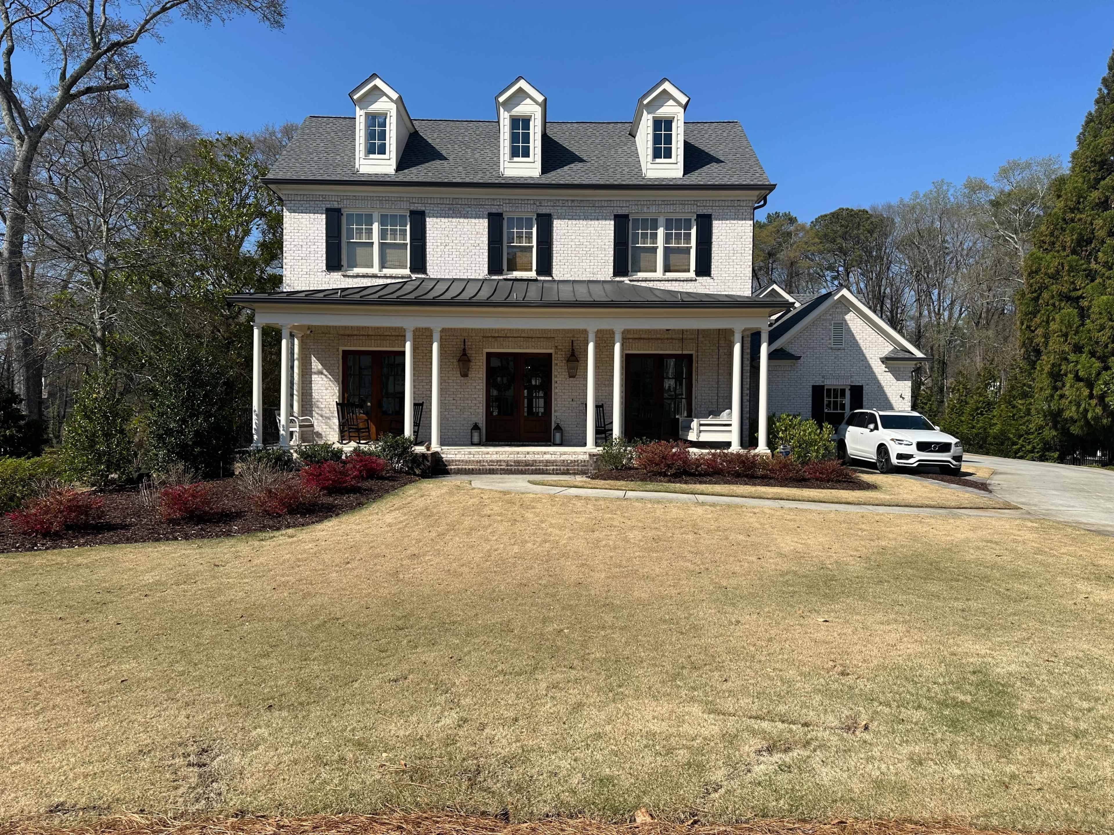 A two-story house with a brick exterior and a gabled roof is surrounded by landscaped greenery and a circular driveway.