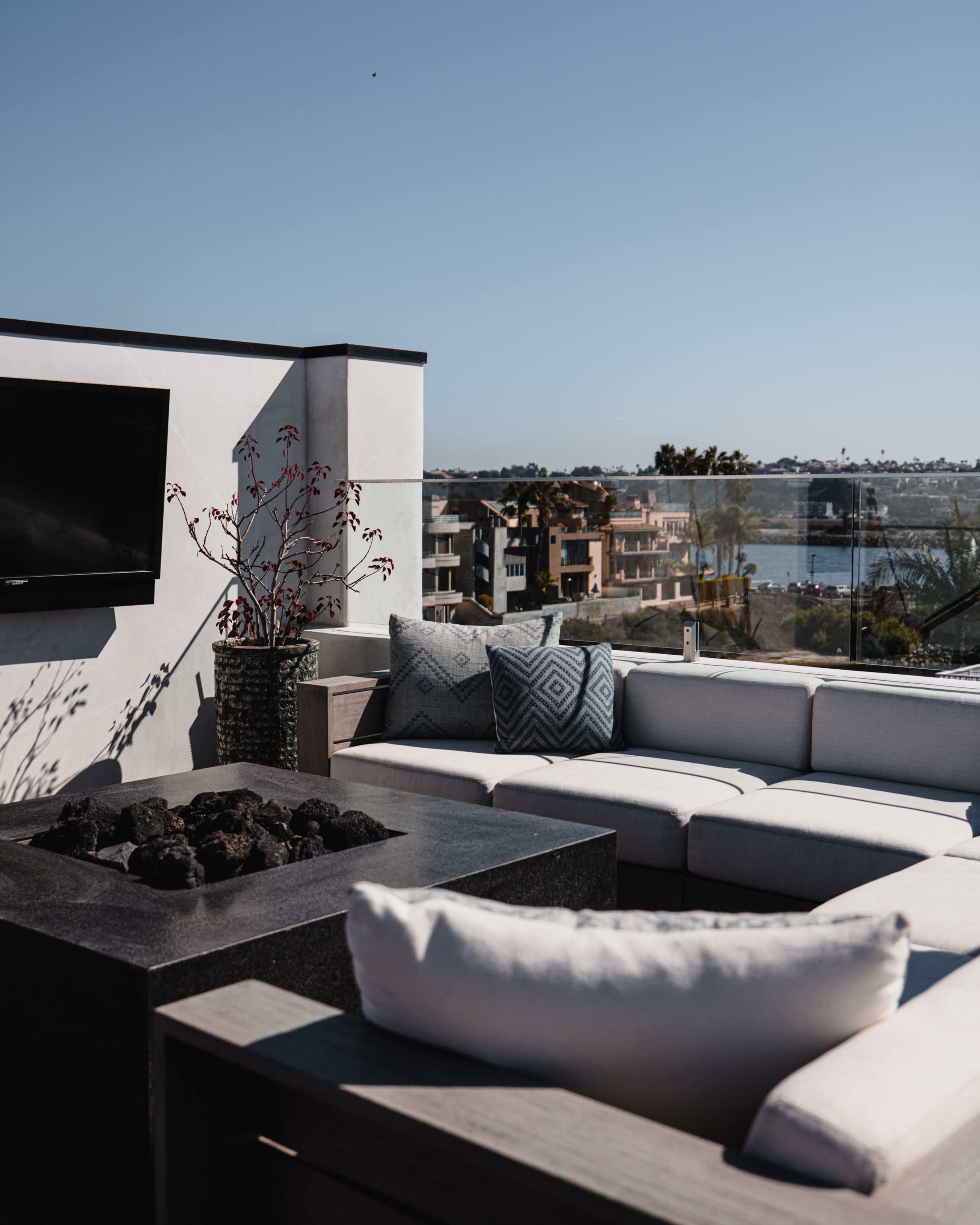 A modern outdoor seating area features a sectional sofa and a fire pit overlooking a body of water and nearby buildings.