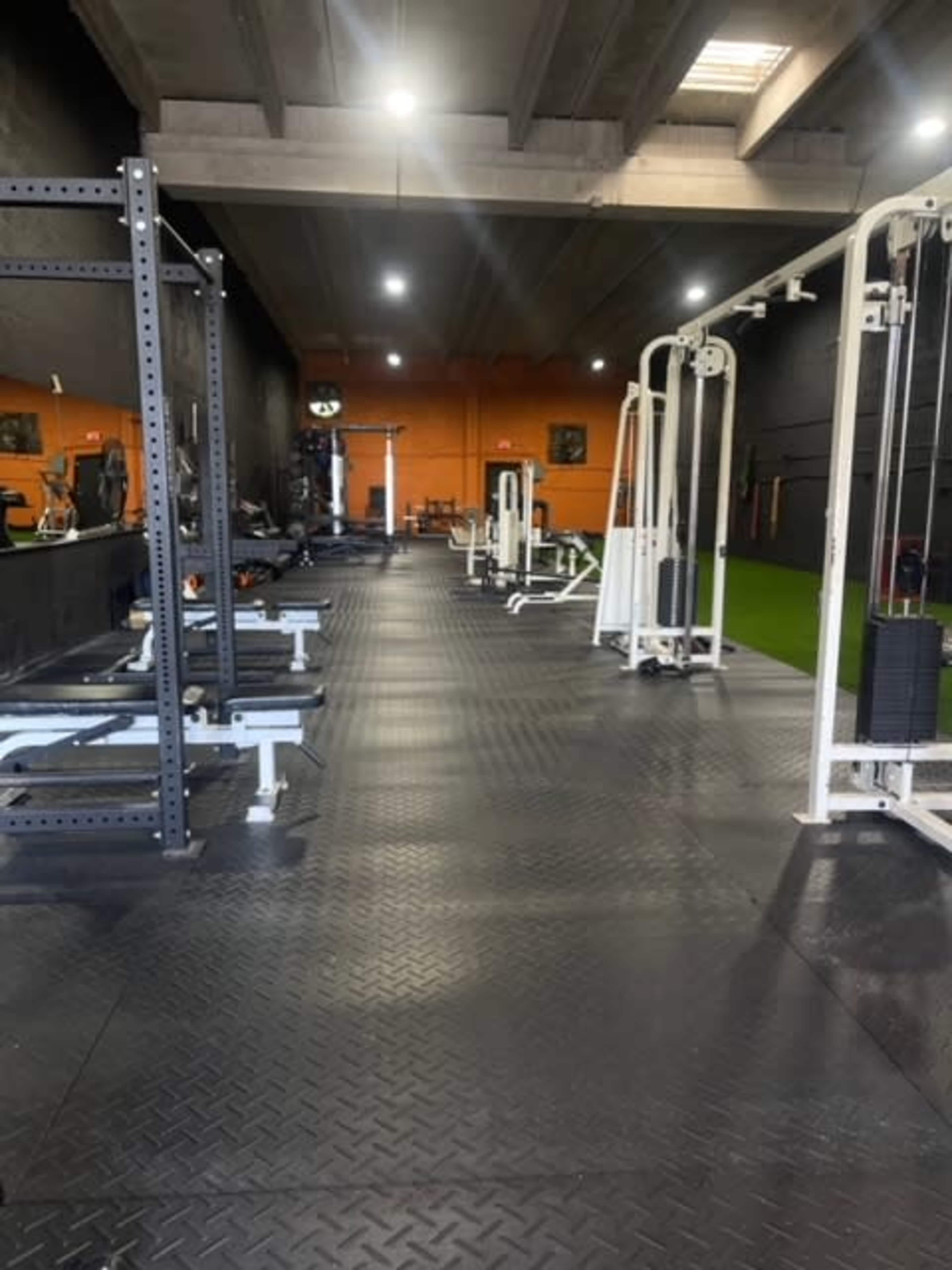 The image shows an empty gym featuring various weightlifting equipment and exercise machines arranged in a long, well-lit space with dark walls and an orange backdrop.