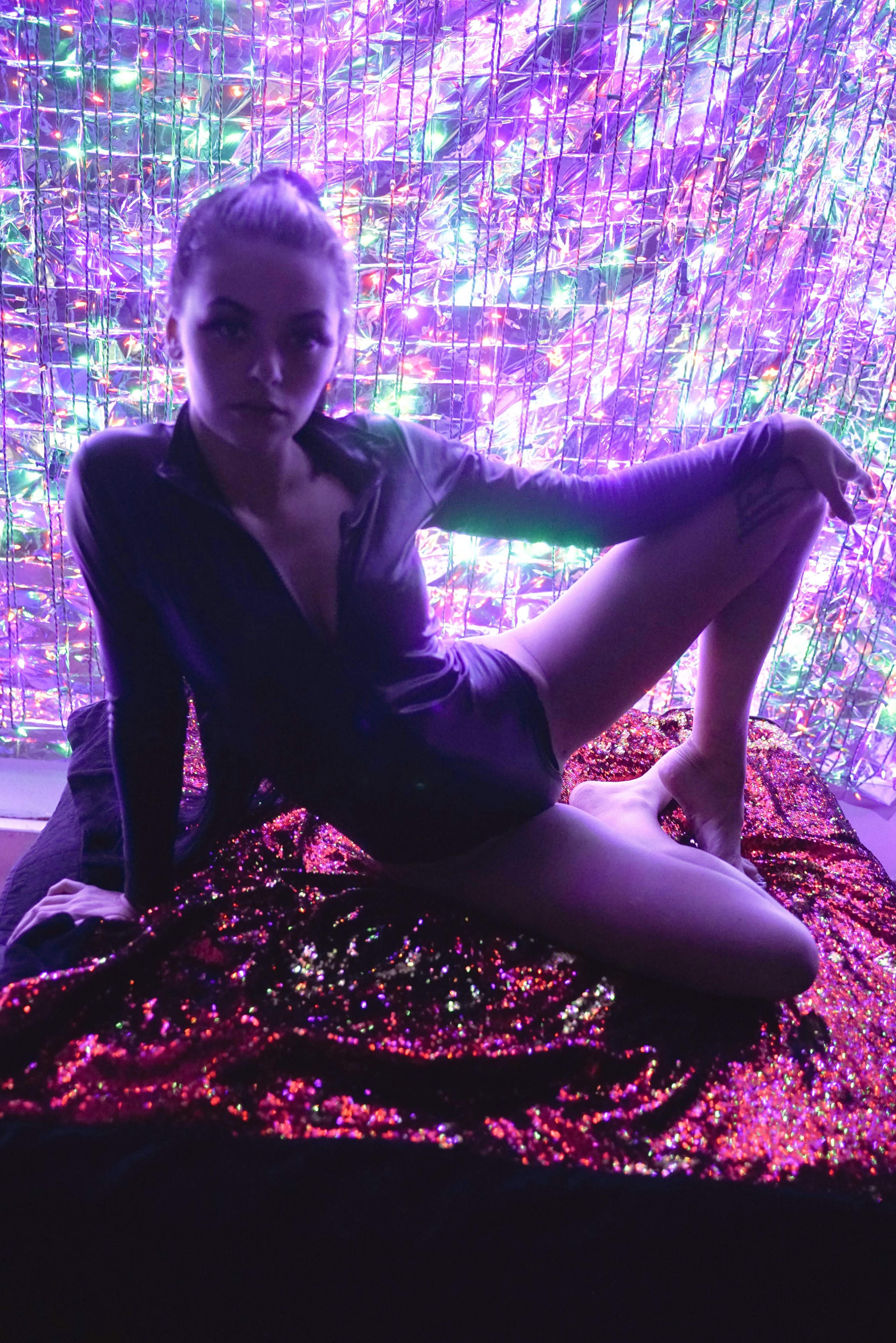 A person wearing a dark outfit poses on a shiny, sequin-covered surface in front of a colorful, lit backdrop.