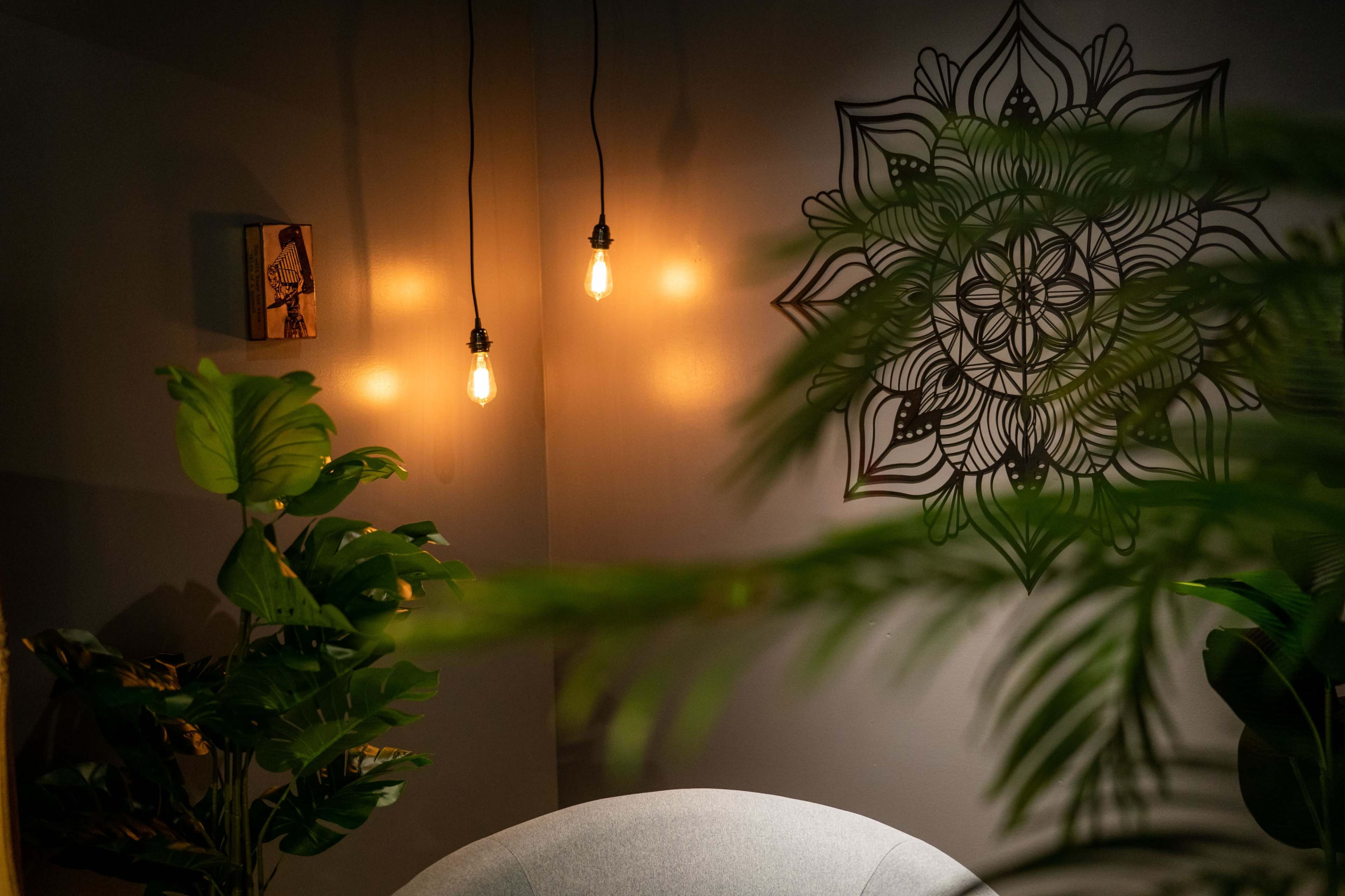 A cozy indoor corner with hanging light bulbs, a decorative wall mandala, and lush green plants surrounding a gray chair.