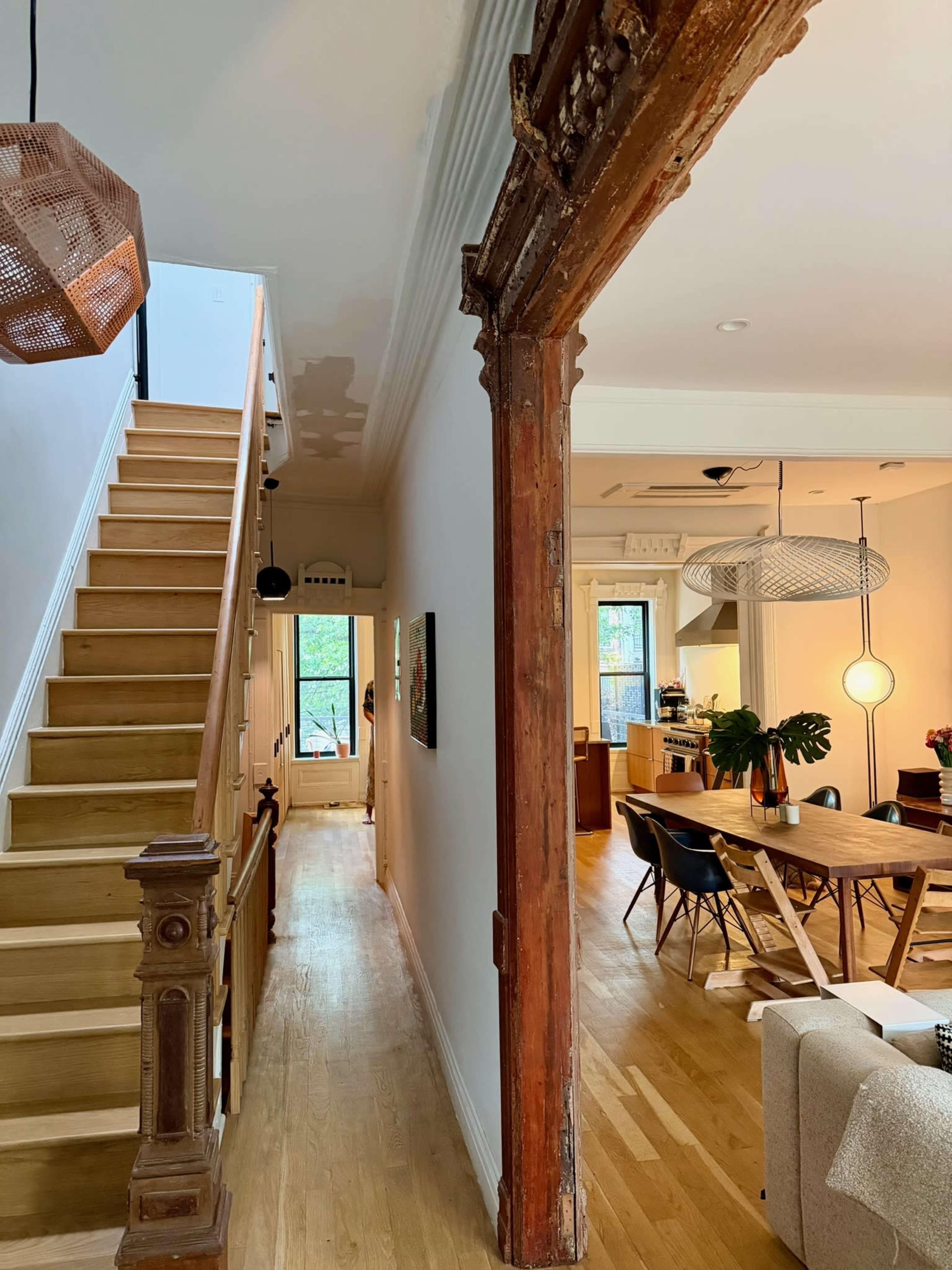 The image shows a hallway in a house featuring a staircase on the left, a visible wood beam supporting the structure, and a dining area with a large table in the background.