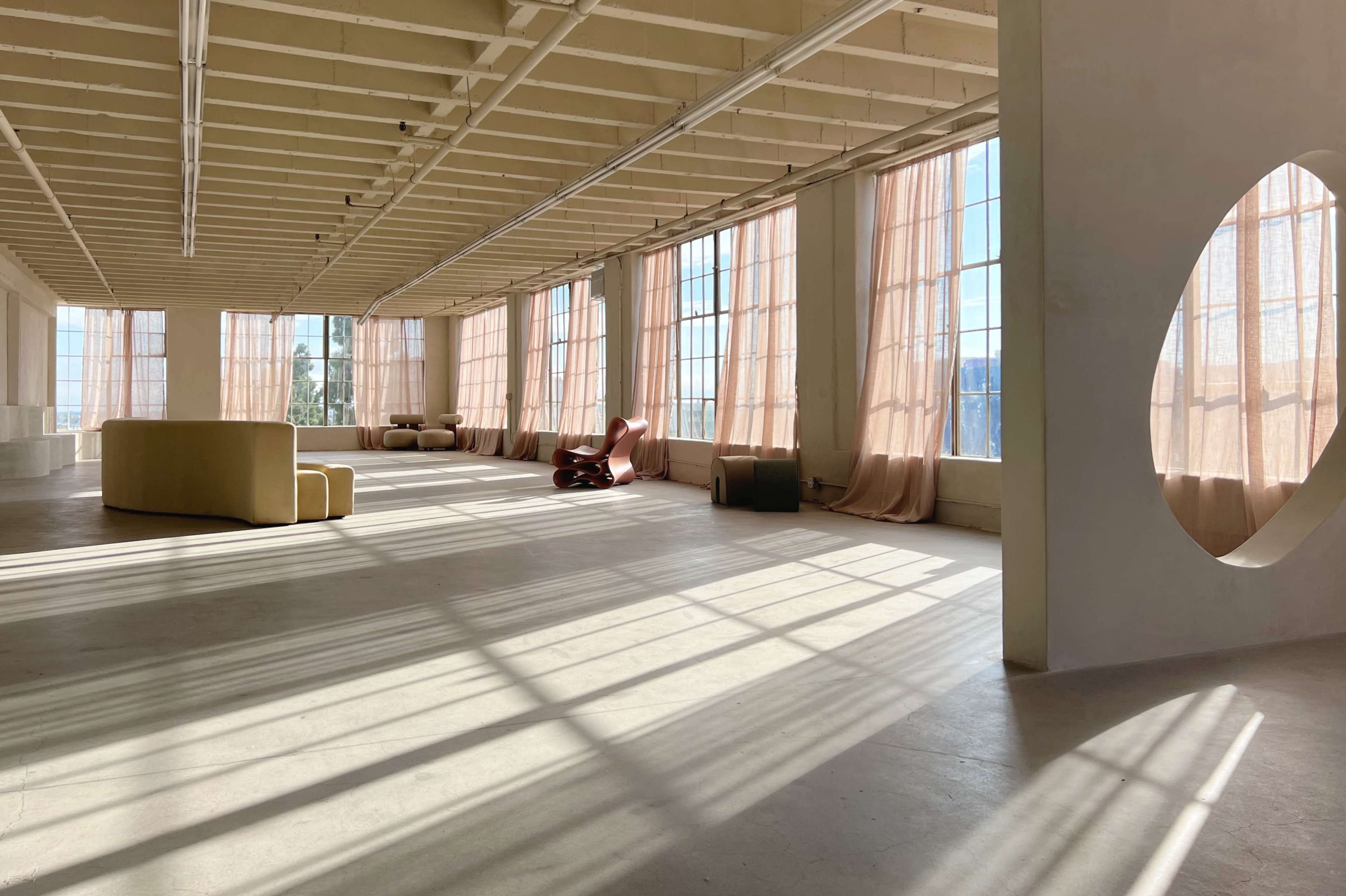 A spacious, sunlit room features large windows with sheer curtains, minimal furniture, and shadow patterns on the floor.