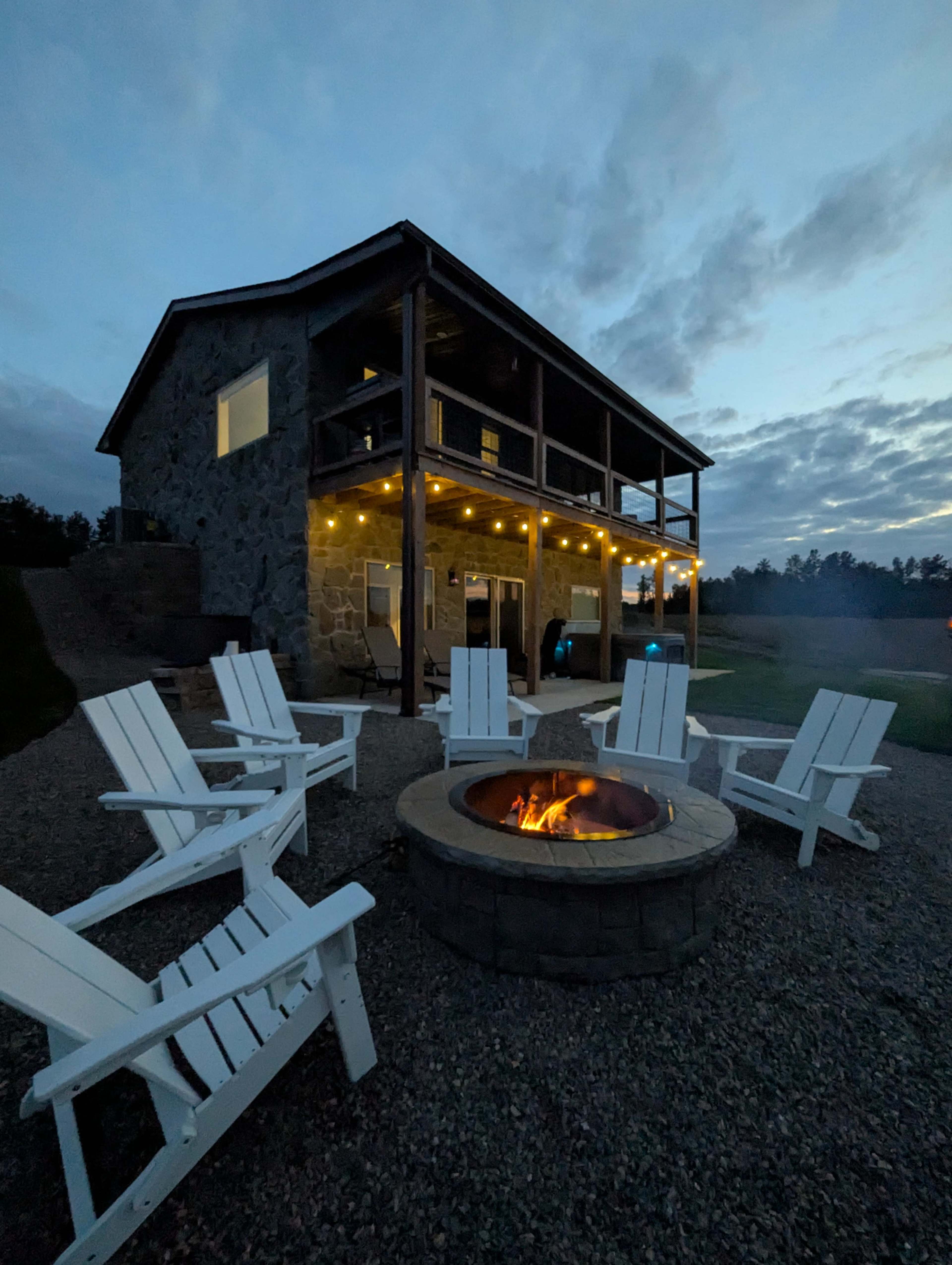 A stone house with a multi-level design features a fire pit surrounded by several white Adirondack chairs, illuminated by string lights in the evening sky.