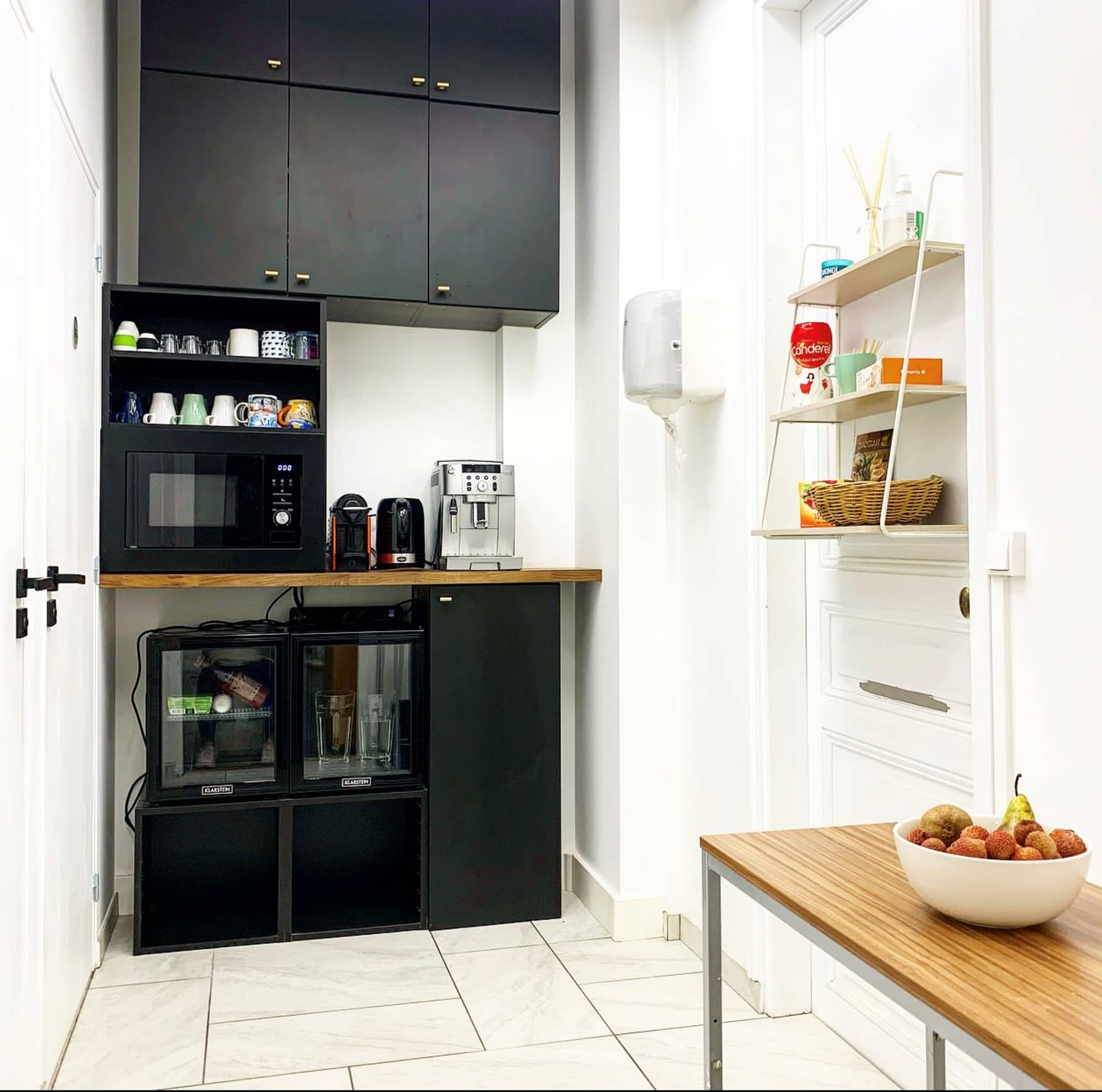 The image shows a modern kitchen corner featuring a black cabinet with a microwave, coffee maker, and a shelf with kitchen items, alongside a small table with a bowl of fruit.