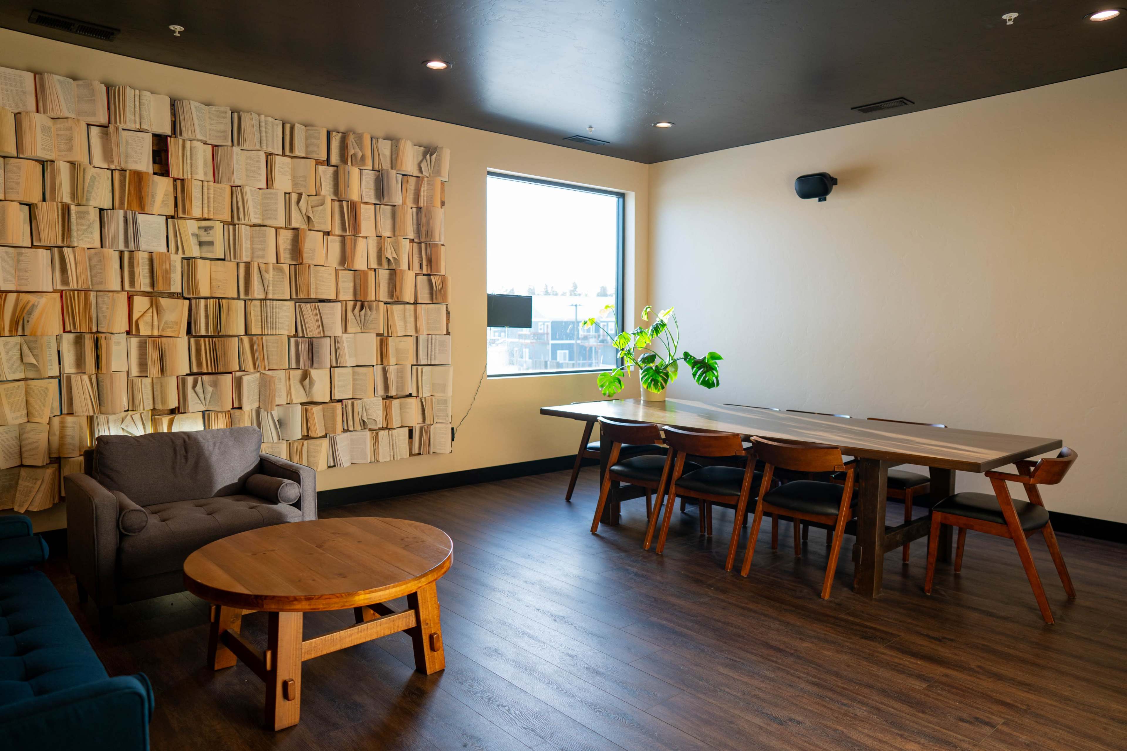 The image shows a modern interior space featuring a wall made of arranged books, a window, a gray armchair, a round wooden coffee table, and a rectangular dining table with chairs.