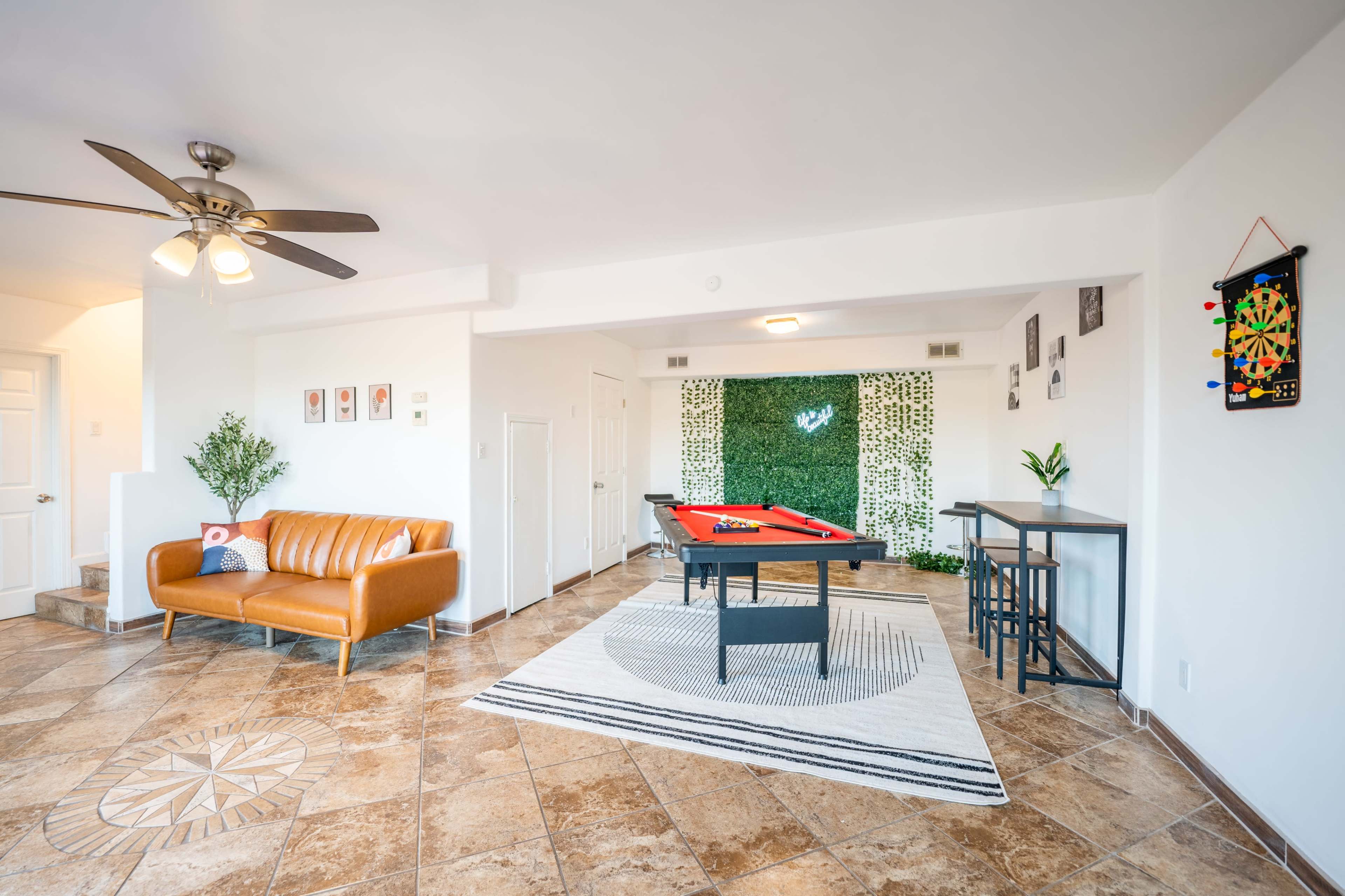 The image shows a well-lit living space with a pool table, a brown leather sofa, and a decorative wall covered in green foliage.
