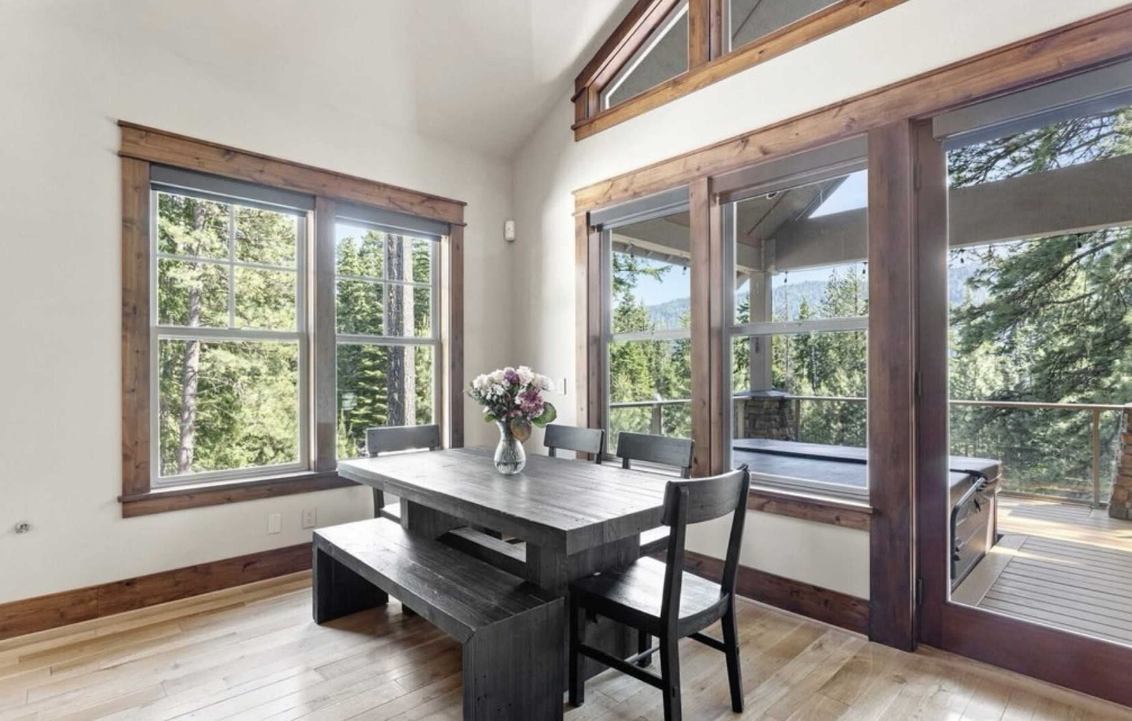 A dining area features a wooden table surrounded by chairs, with large windows offering views of a forest outside.