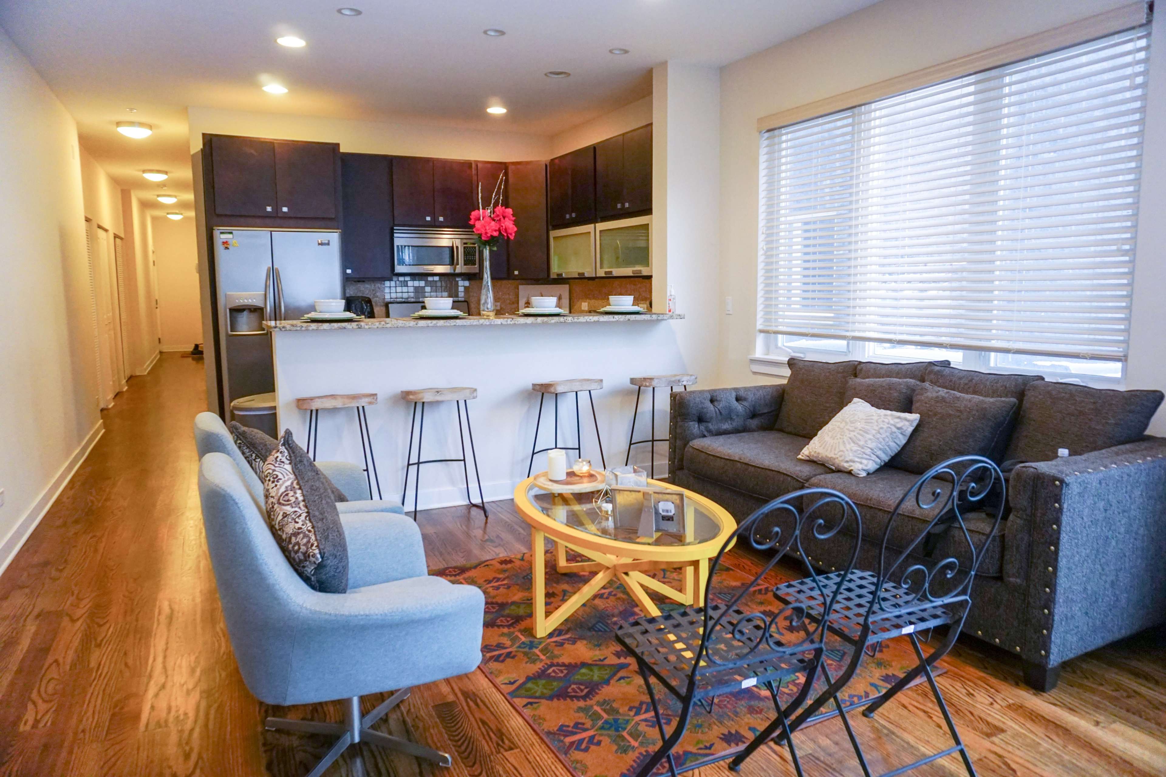 A modern living space features a kitchen bar with stools, a gray sofa, a yellow coffee table, and two outdoor chairs, set against a backdrop of light-colored walls and a hallway.