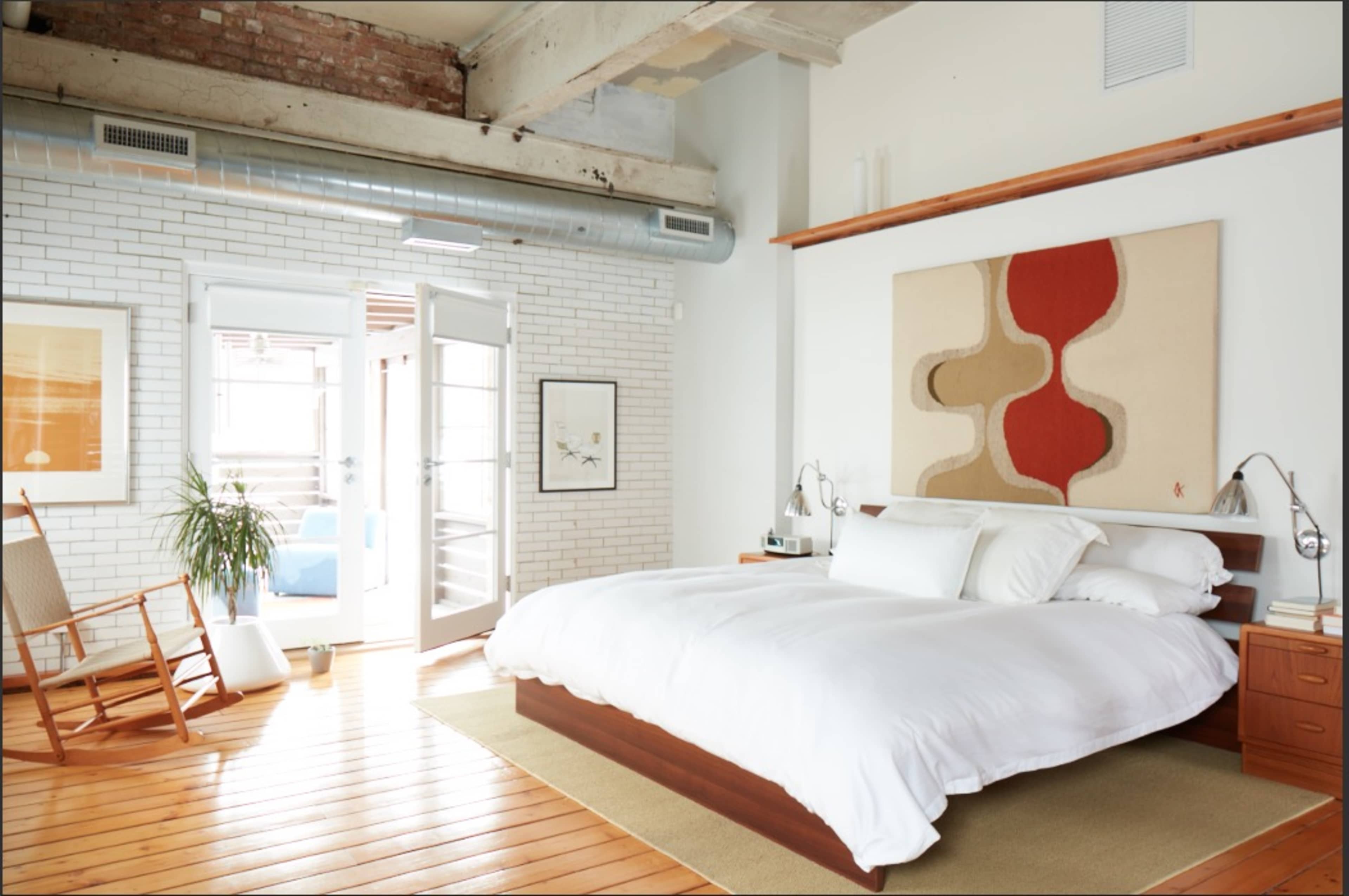 The image shows a spacious bedroom featuring a large bed with white bedding, a wooden nightstand, a rocking chair, and art pieces on the walls, with double doors leading to an outdoor space.