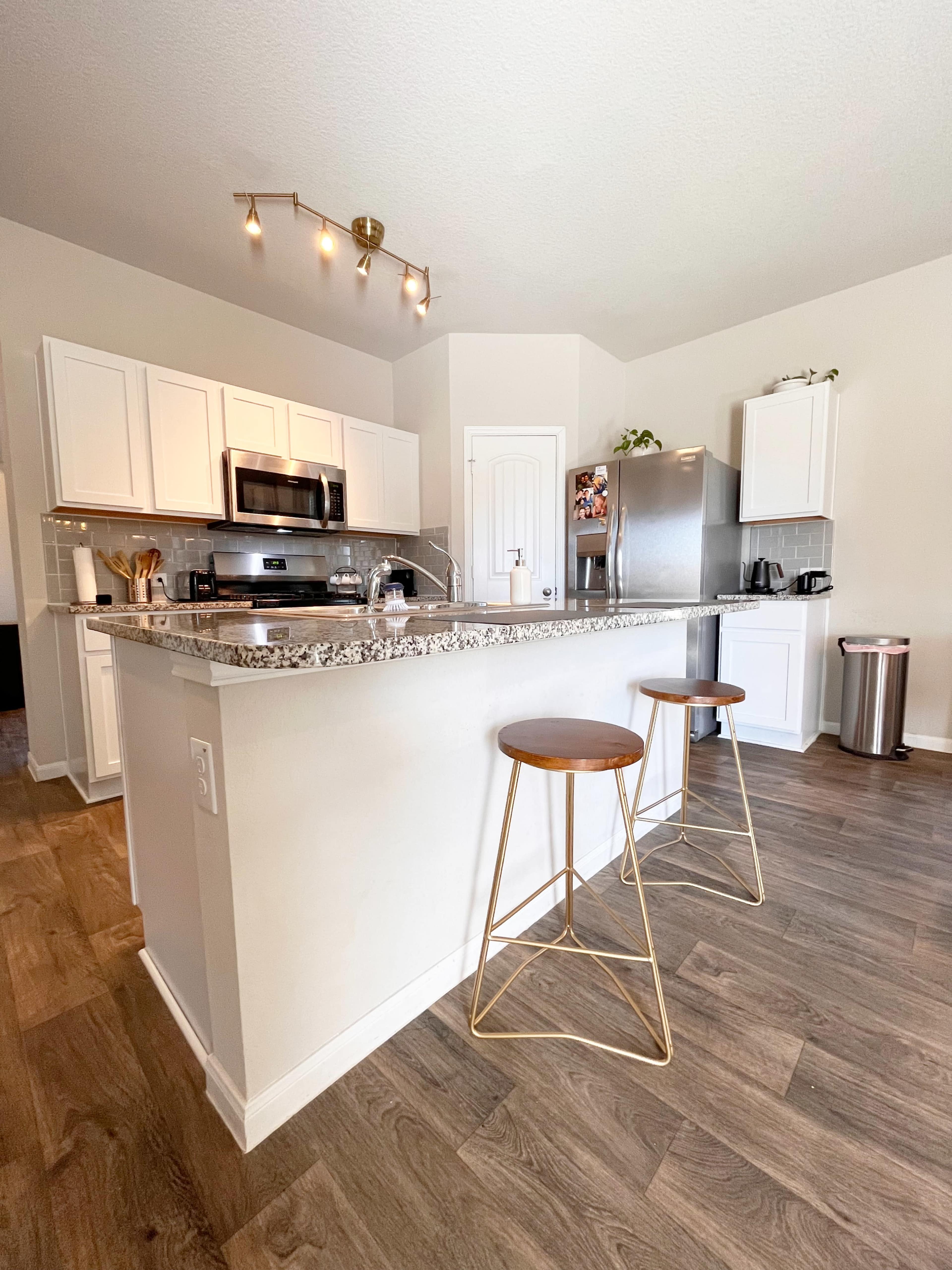 A modern kitchen features white cabinets, stainless steel appliances, and a granite-topped island with two gold bar stools.