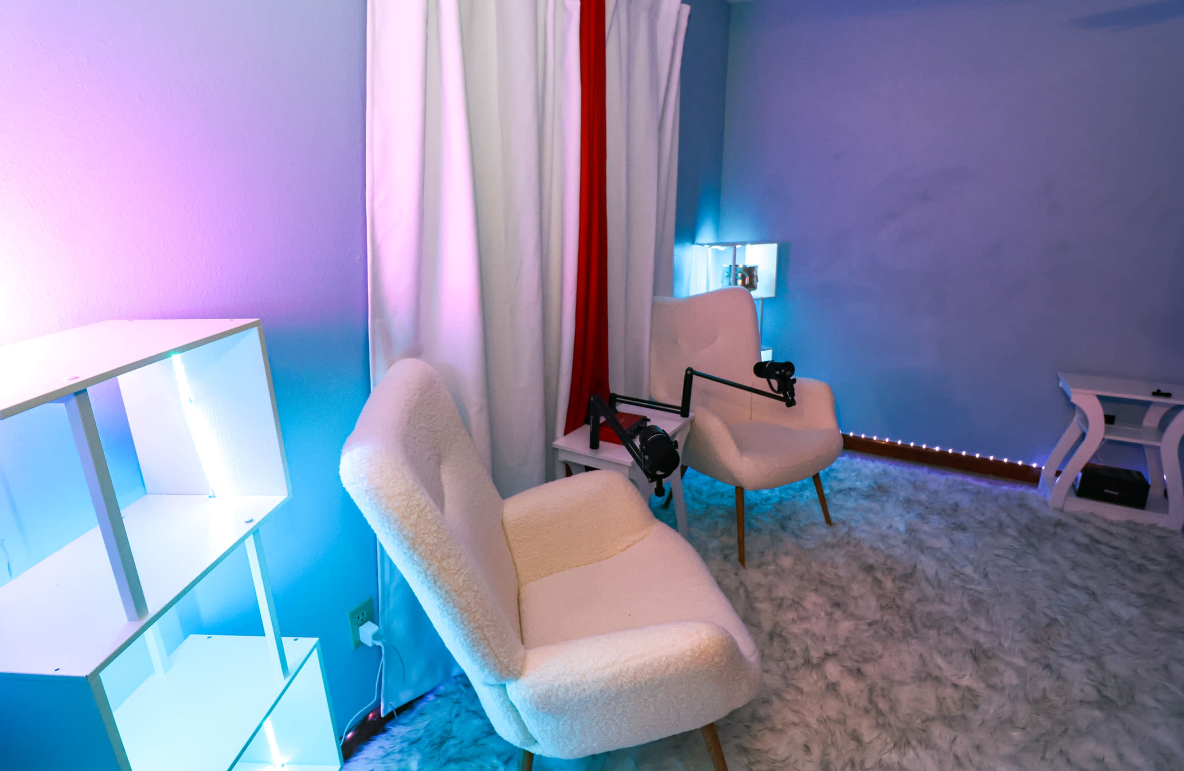 The image shows a softly lit room with two white chairs on a fluffy carpet, accompanied by a shelf and a microphone setup.