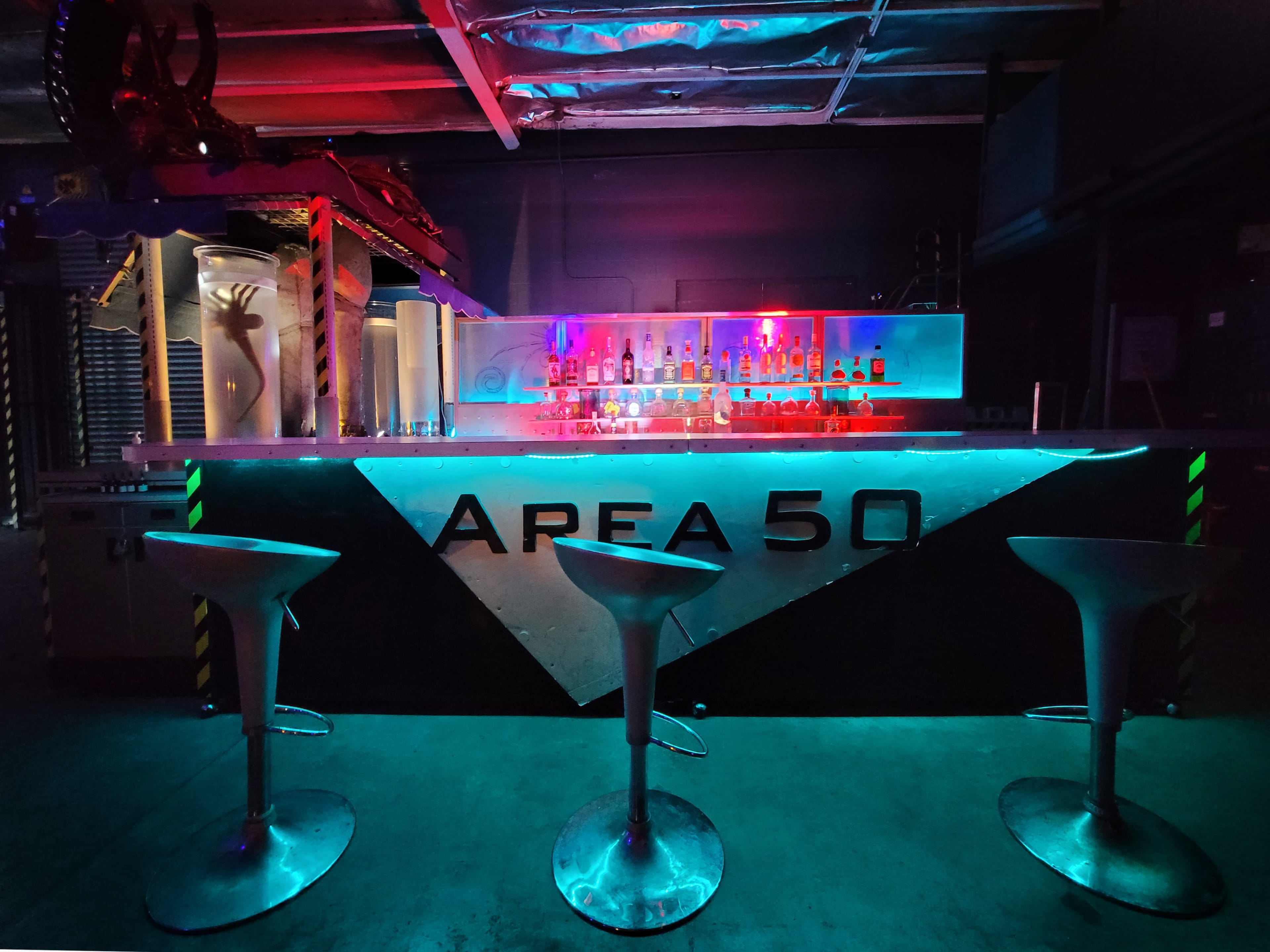 A brightly lit bar with a futuristic design, featuring the name "AREA 50" prominently displayed and accompanied by three metallic stools.