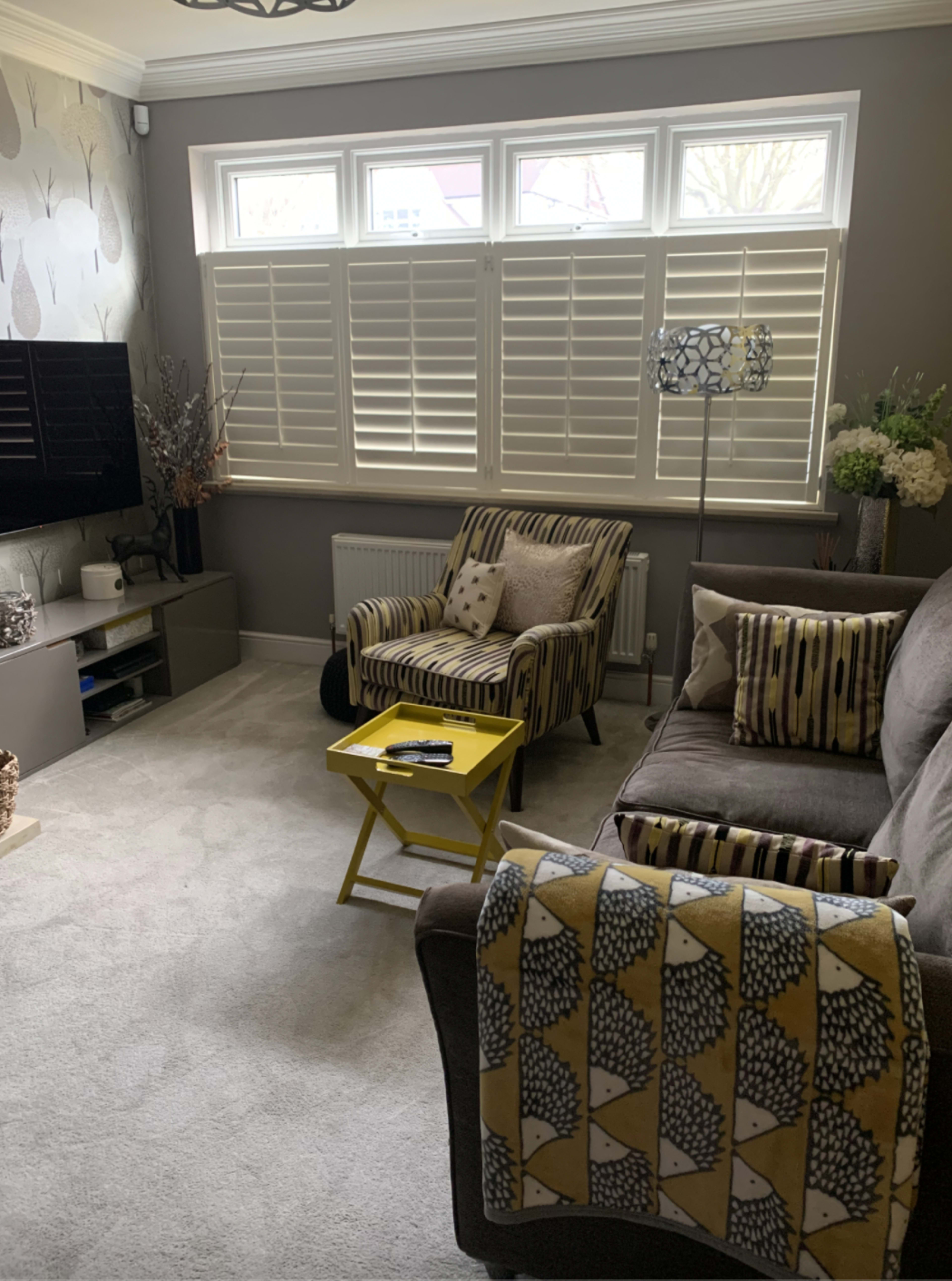 The living room features a gray sofa and an armchair with patterned cushions, alongside a small yellow table, a television, and large shuttered windows.