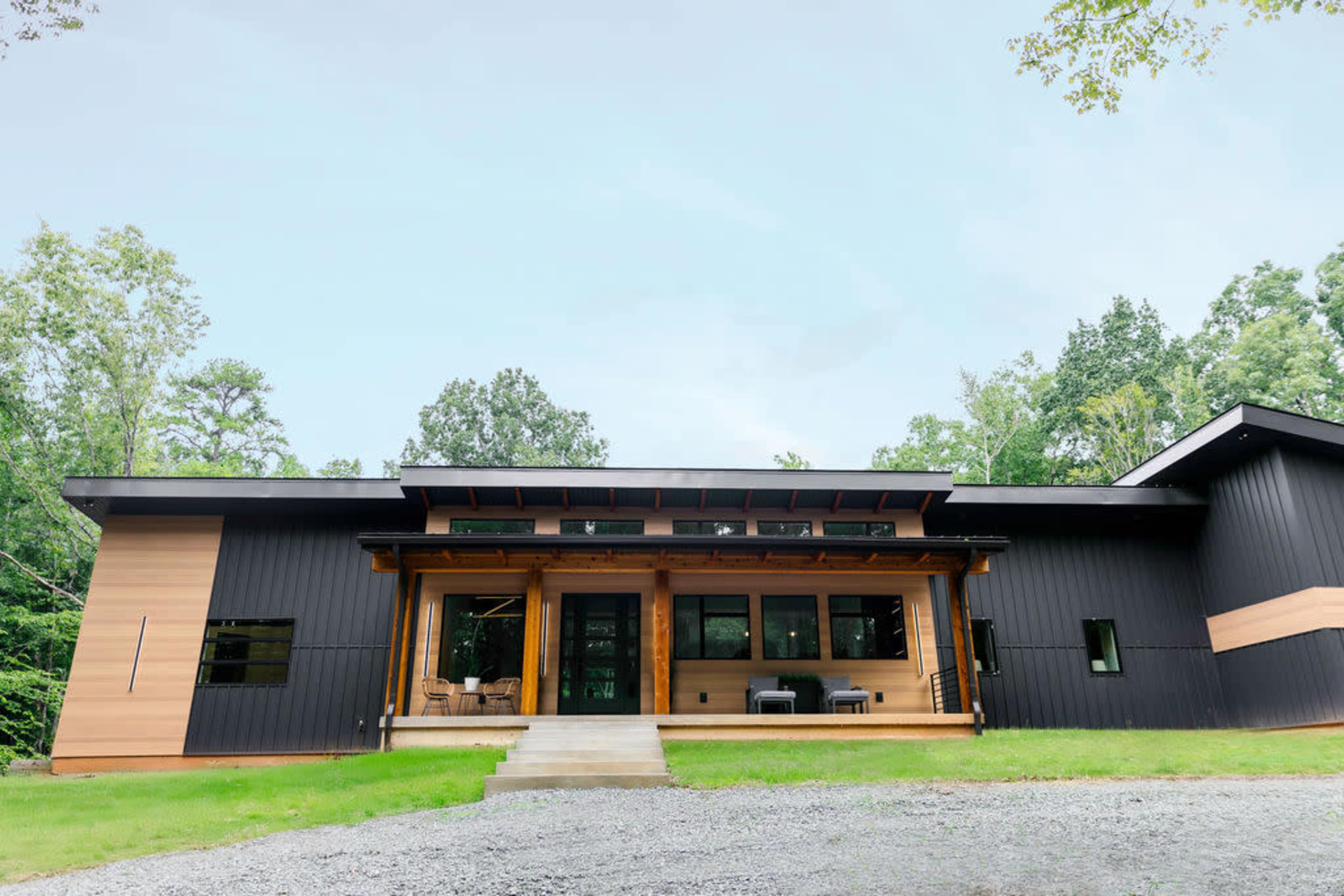 A modern house features a combination of dark wood and metal siding, with large windows and a covered porch surrounded by greenery.