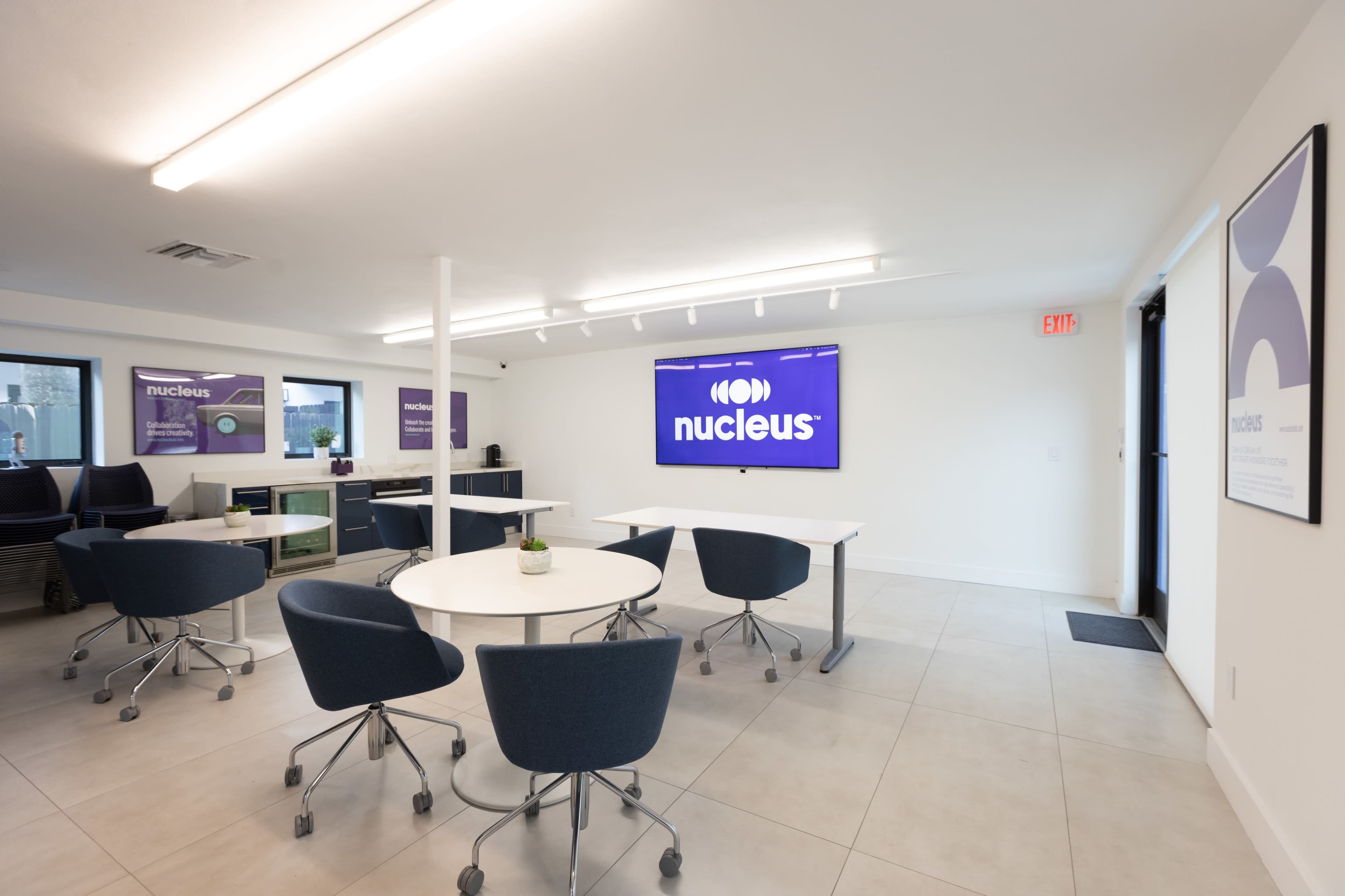 A modern office space featuring tables and chairs, a large screen displaying the word "nucleus," and purple-themed wall art.