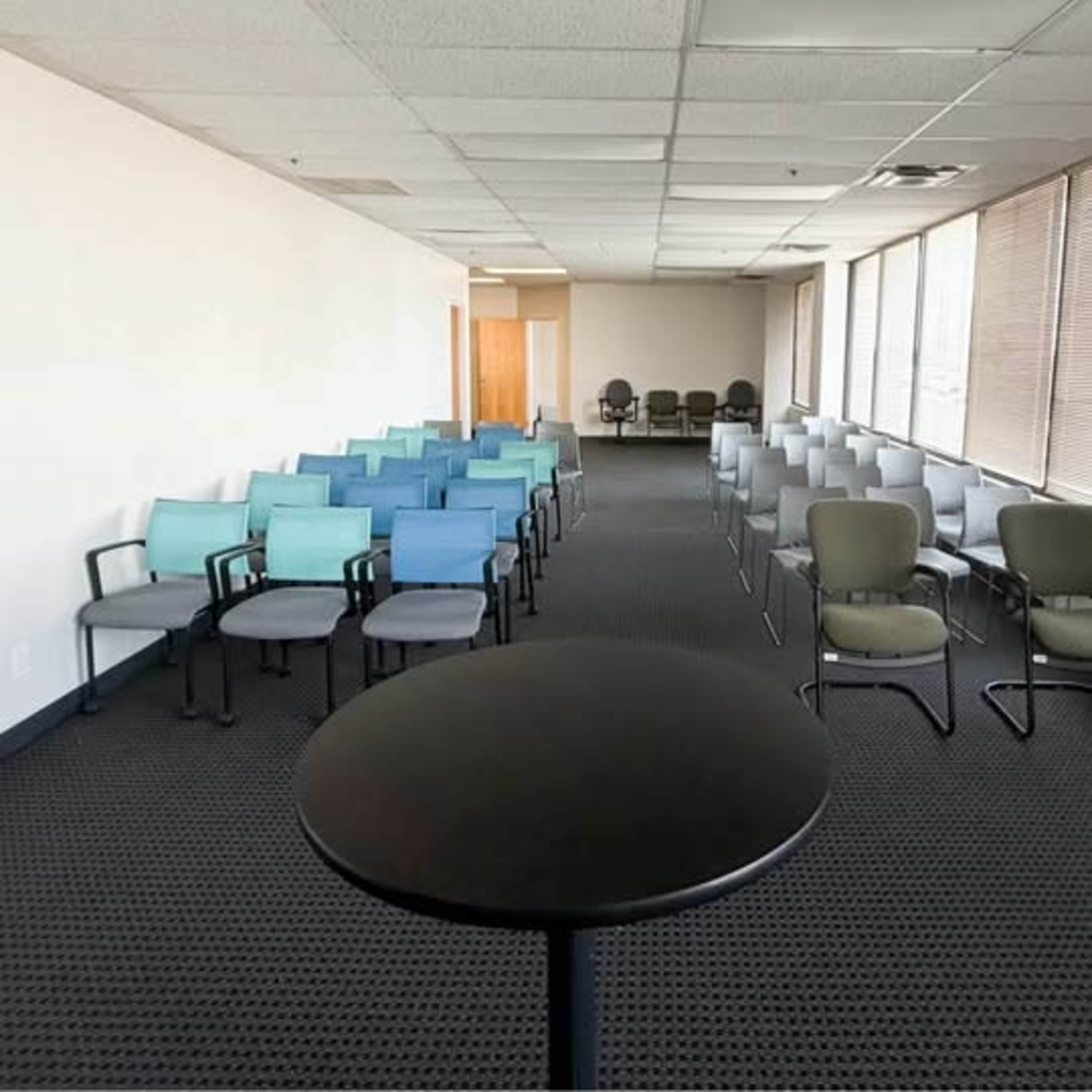 The image shows a large, empty room with rows of chairs arranged in two different colors and a small black table in the foreground.