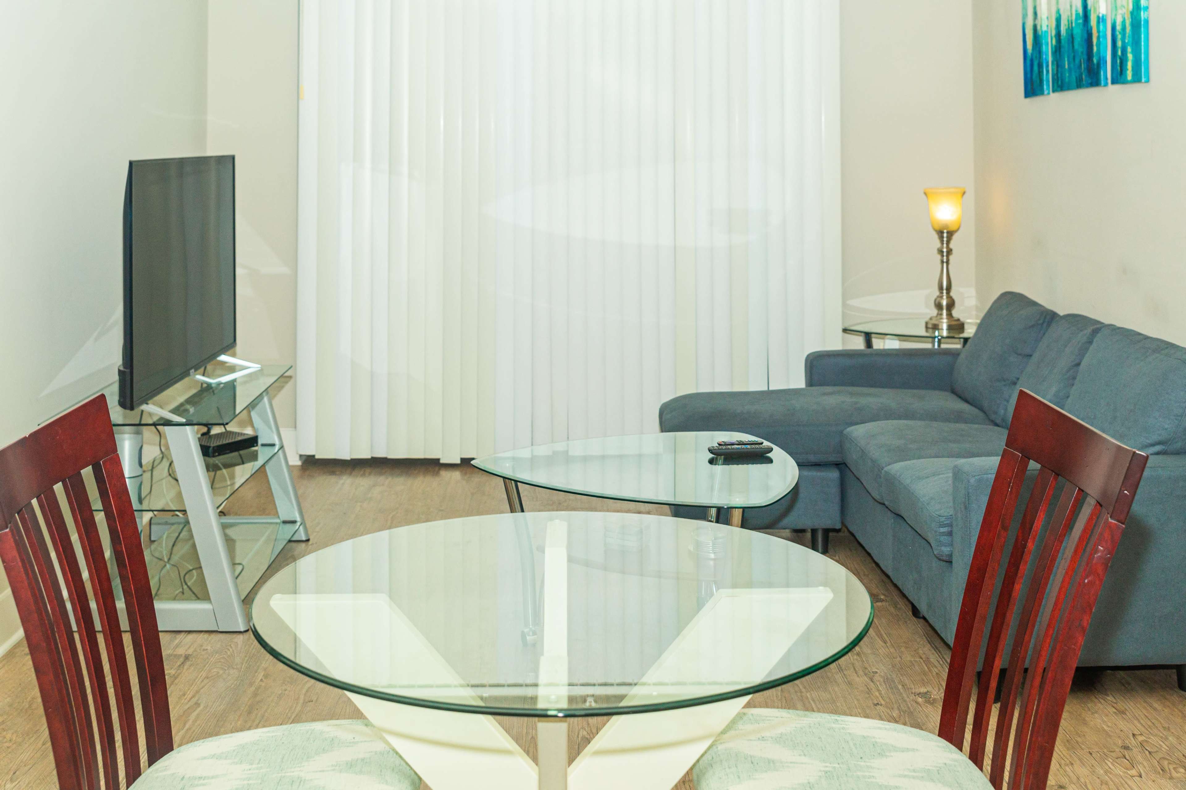 A modern living room features a glass dining table, a gray sectional sofa, a television on a stand, and vertical blinds covering the window.