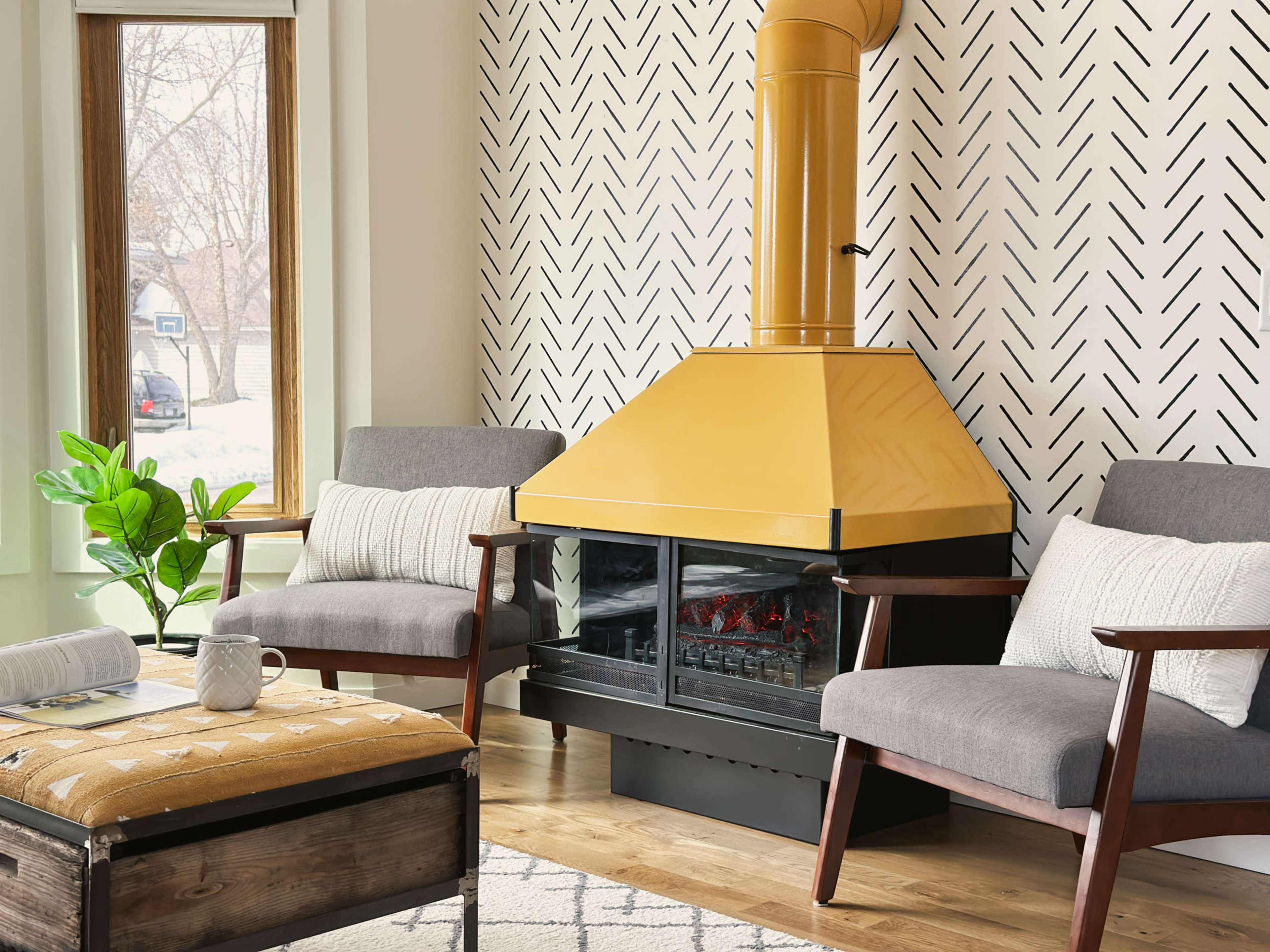 A living room features two gray chairs flanking a yellow triangular fireplace, with a patterned wall and a window revealing an outdoor view.