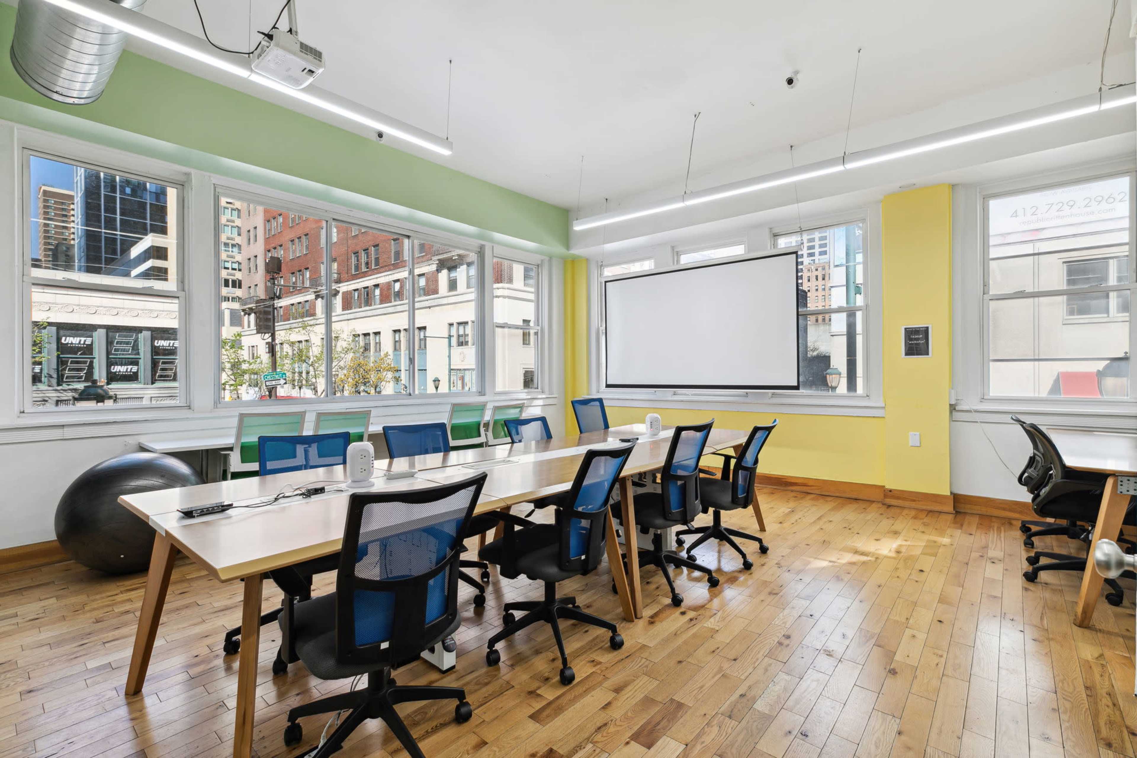 A modern, open office space features a long table with several chairs arranged around it, large windows allowing natural light, and a projector screen on the wall.