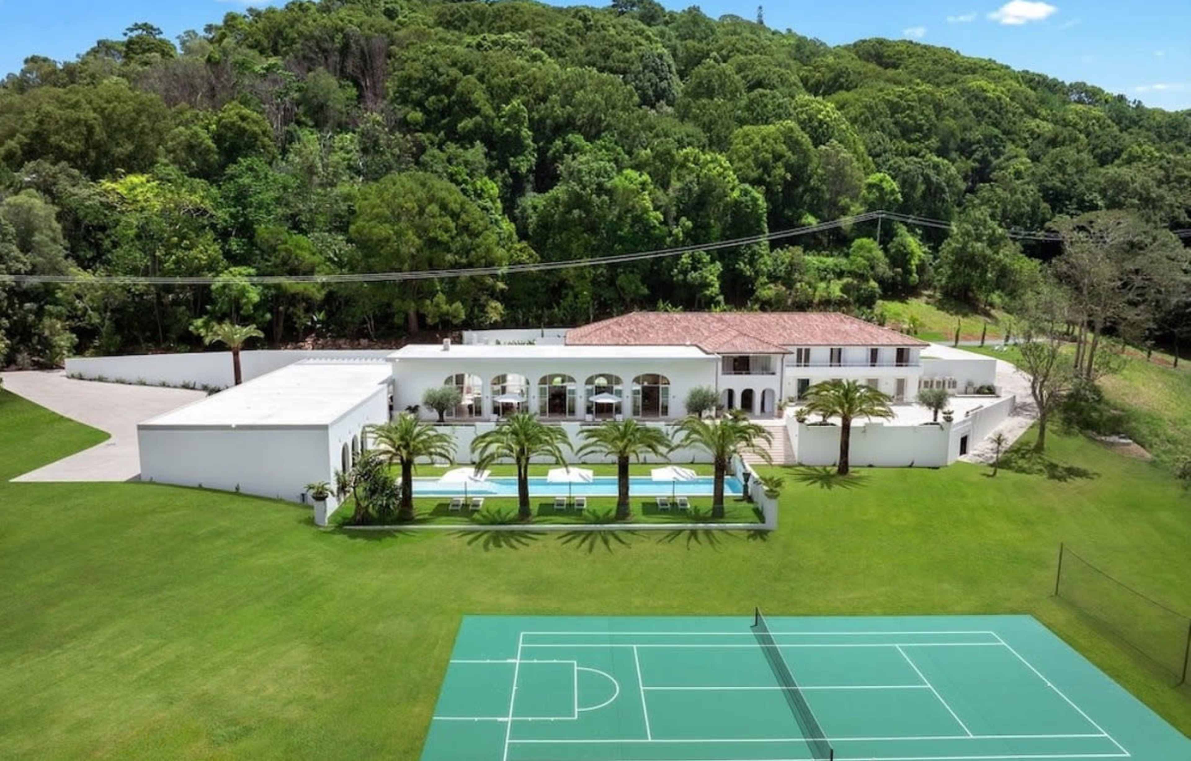 The image shows a large white mansion surrounded by green lawn and trees, with a swimming pool and a tennis court visible in the foreground.