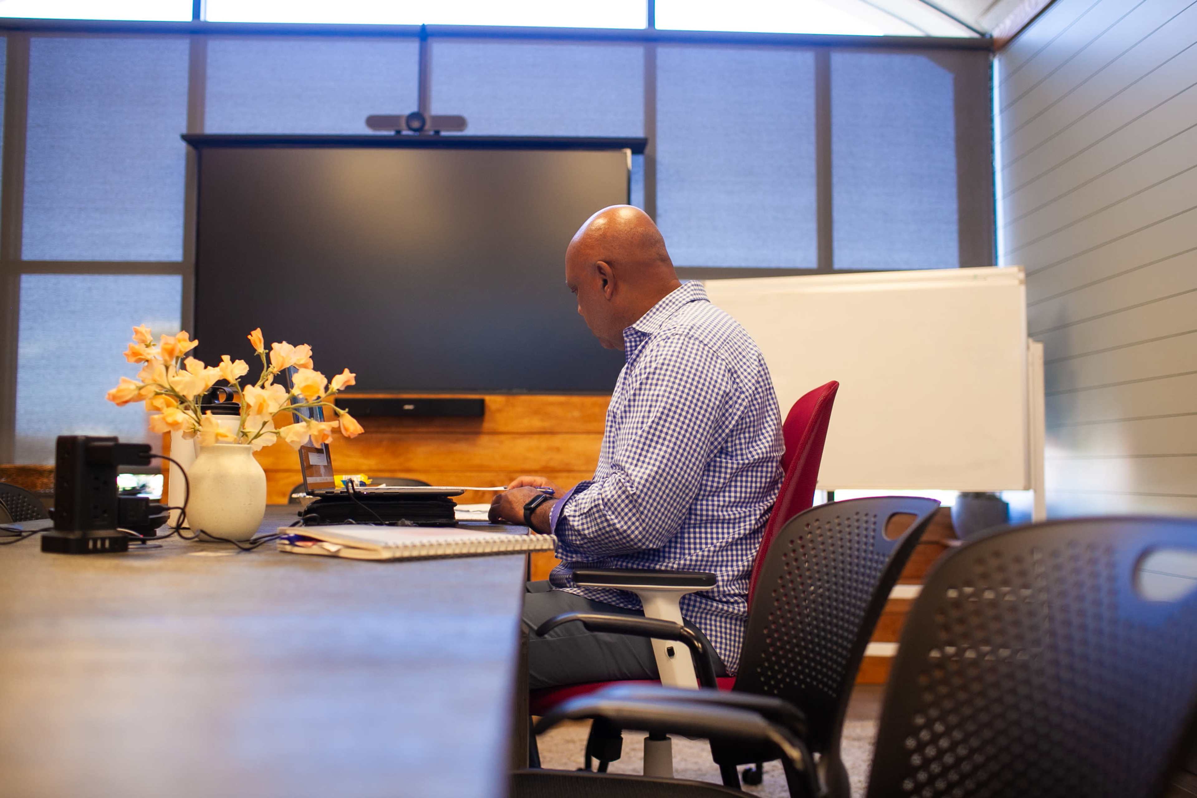 A man in a blue checkered shirt sits at a conference table, working on a laptop with a vase of flowers beside him in a modern meeting room.