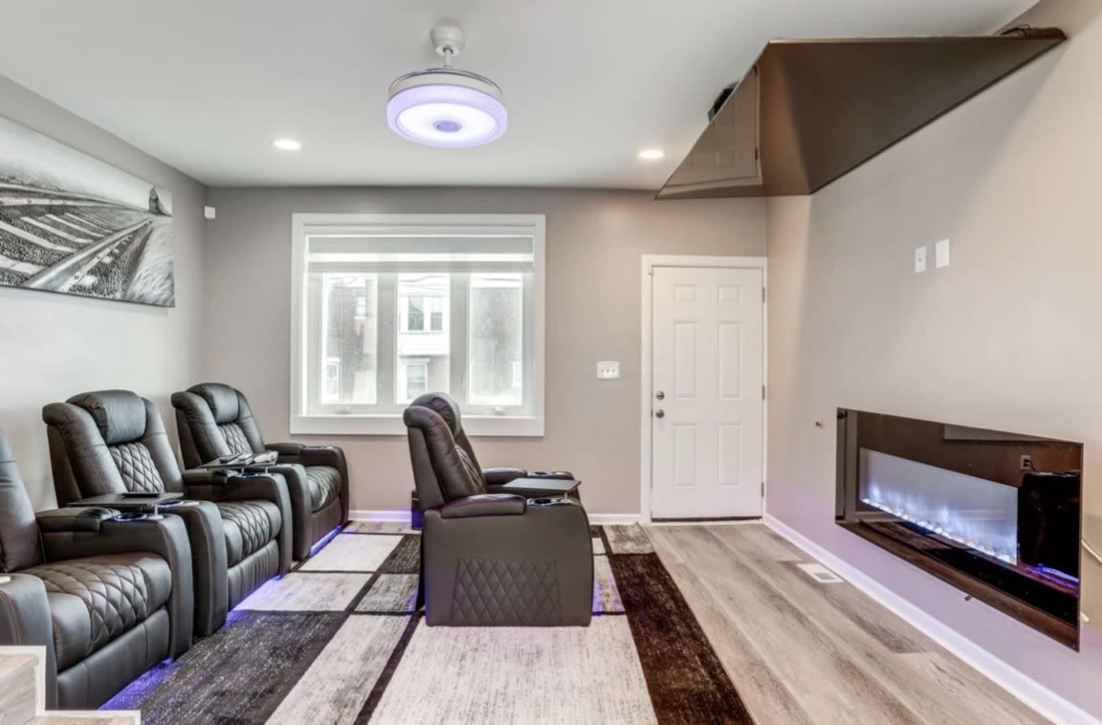 The image shows a modern home theater room featuring three reclining chairs, a sleek electric fireplace, and large windows illuminated by natural light.